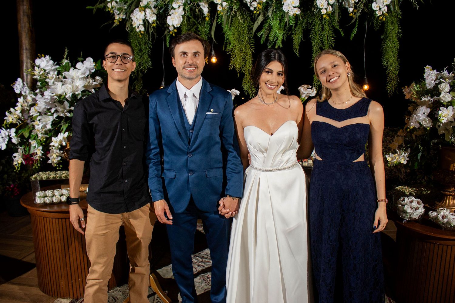 A bride and groom pose with two guests at an elegant wedding ceremony decorated with white flowers and greenery.