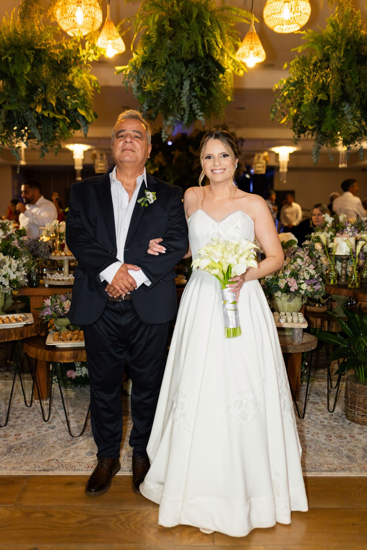 Bride in white gown holding bouquet stands with older man in black suit at elegant wedding reception.