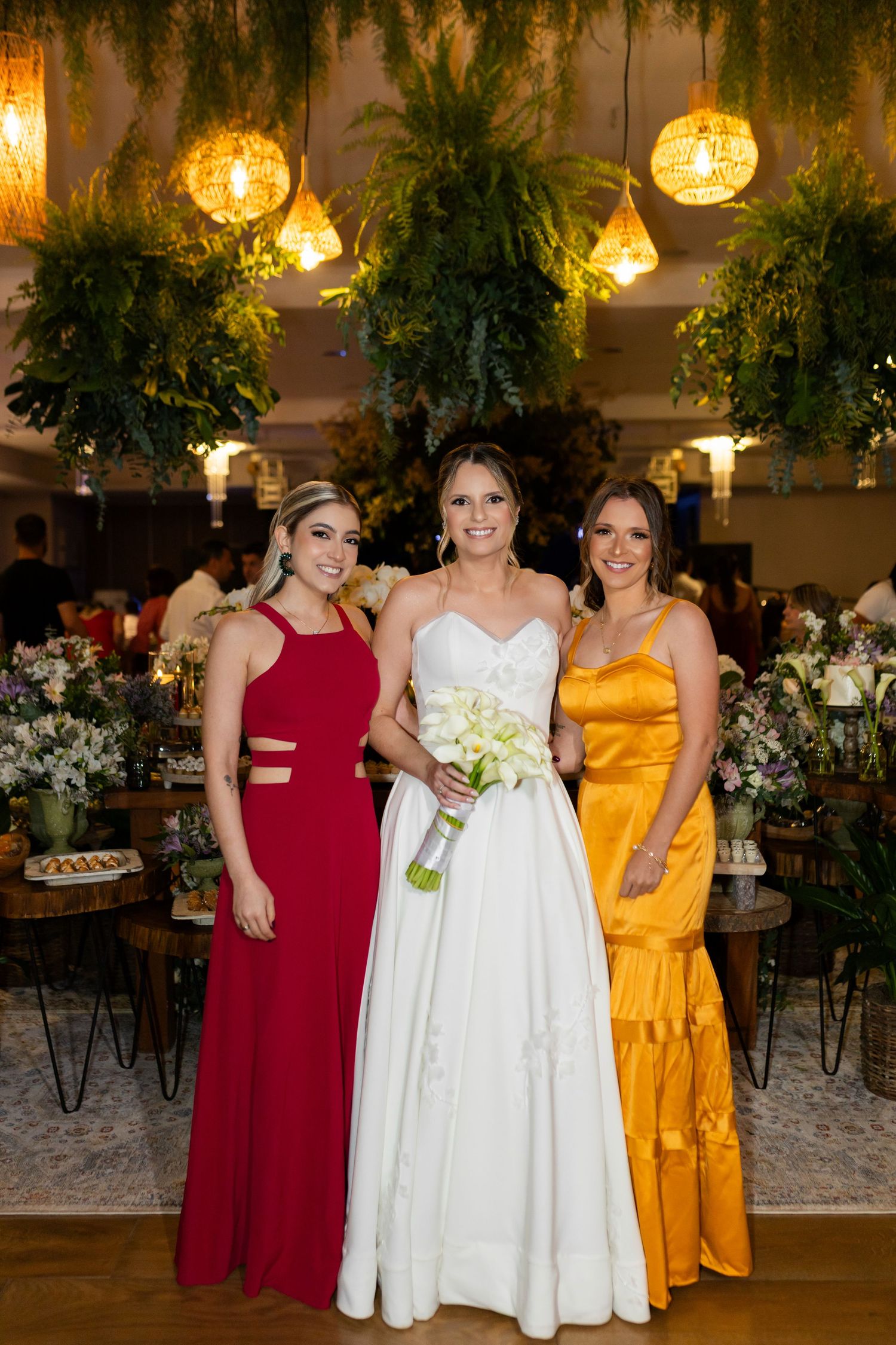 Bride in white gown holds bouquet flanked by bridesmaids in red and yellow dresses at elegant floral wedding venue.