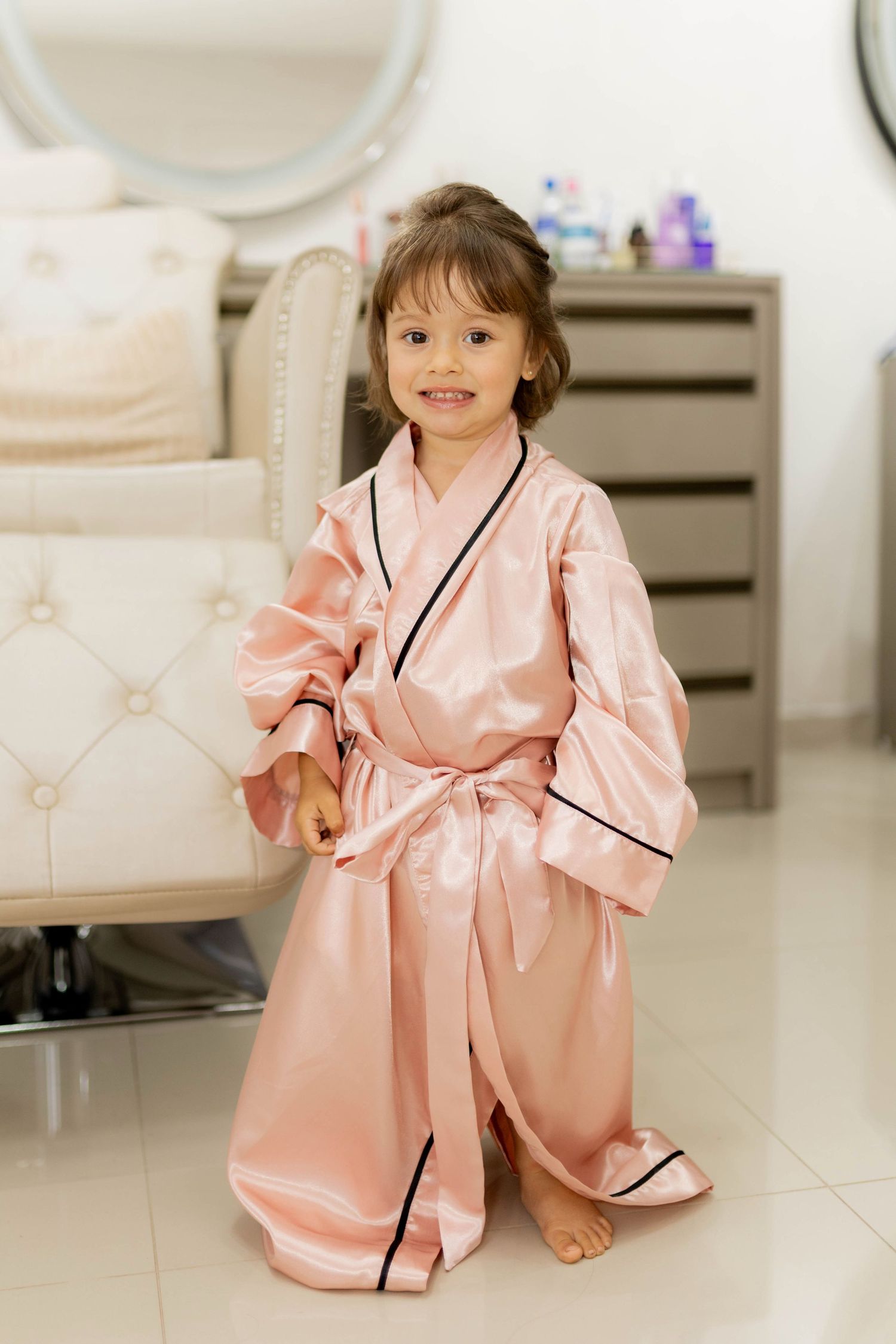 A smiling little girl wearing an oversized pink satin robe with black trim, standing barefoot in a beauty salon.