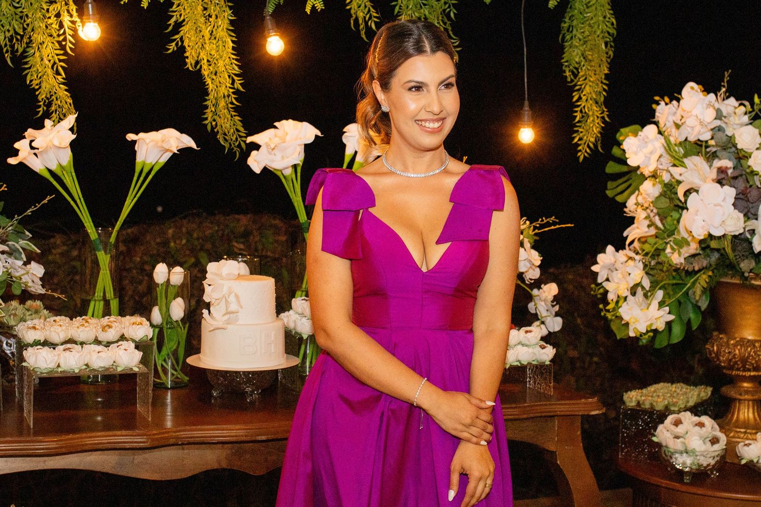 Woman in elegant magenta gown poses at decorated event with white floral arrangements and wedding cake.
