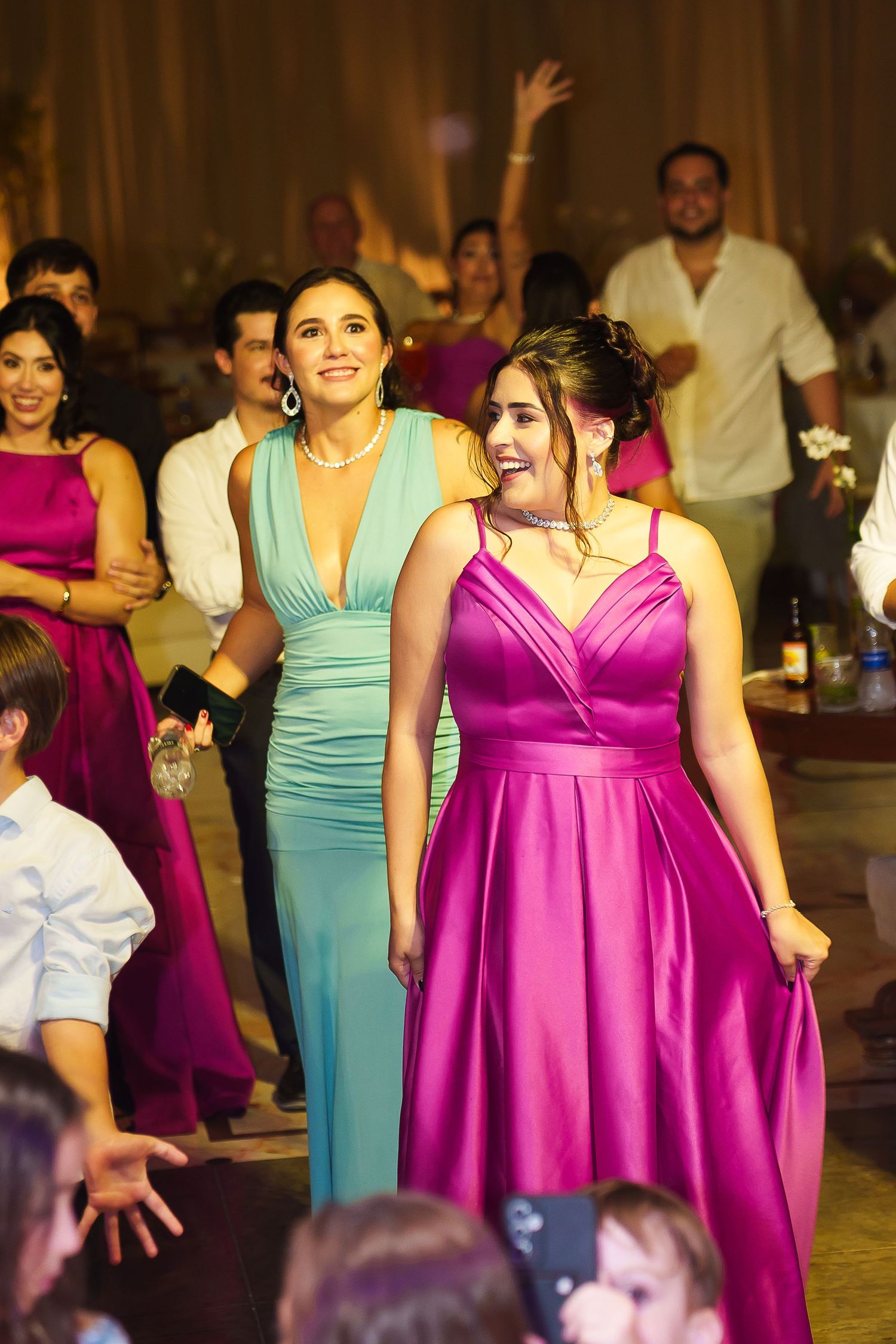 Two women in elegant teal and magenta gowns smile at a festive wedding reception celebration.