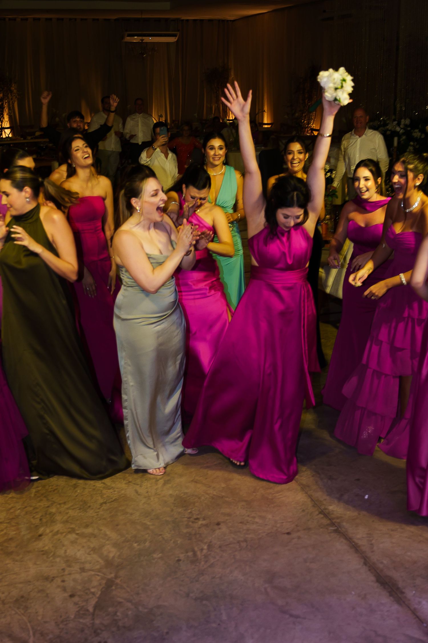 Bridesmaids in magenta and silver gowns celebrate at a wedding reception, with one raising a bouquet in triumph.