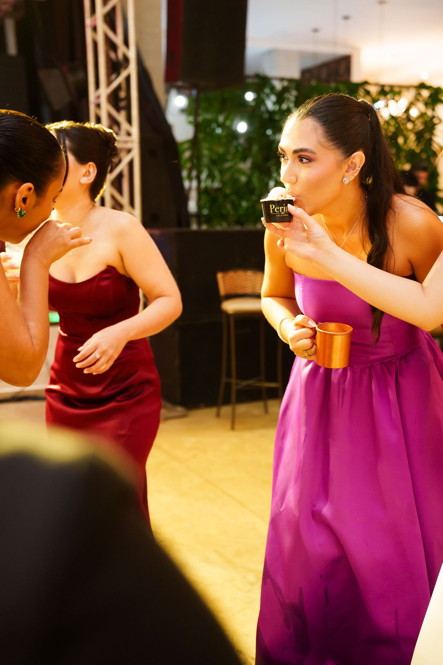 Women in elegant gowns socializing at a formal event, one in purple dress drinking from a cup.