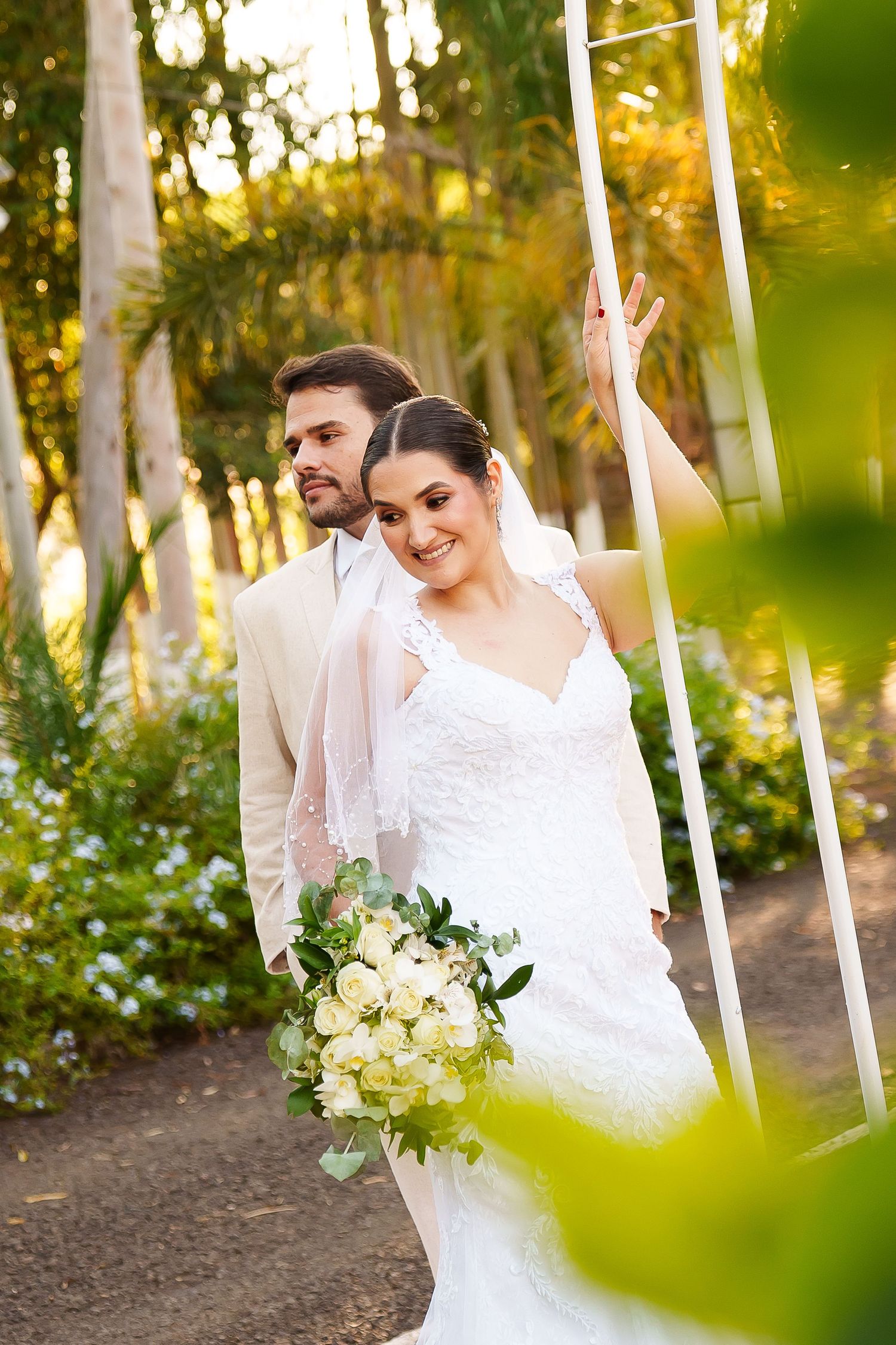 Bride in white lace gown holding bouquet stands with groom in beige suit outdoors among lush green trees.