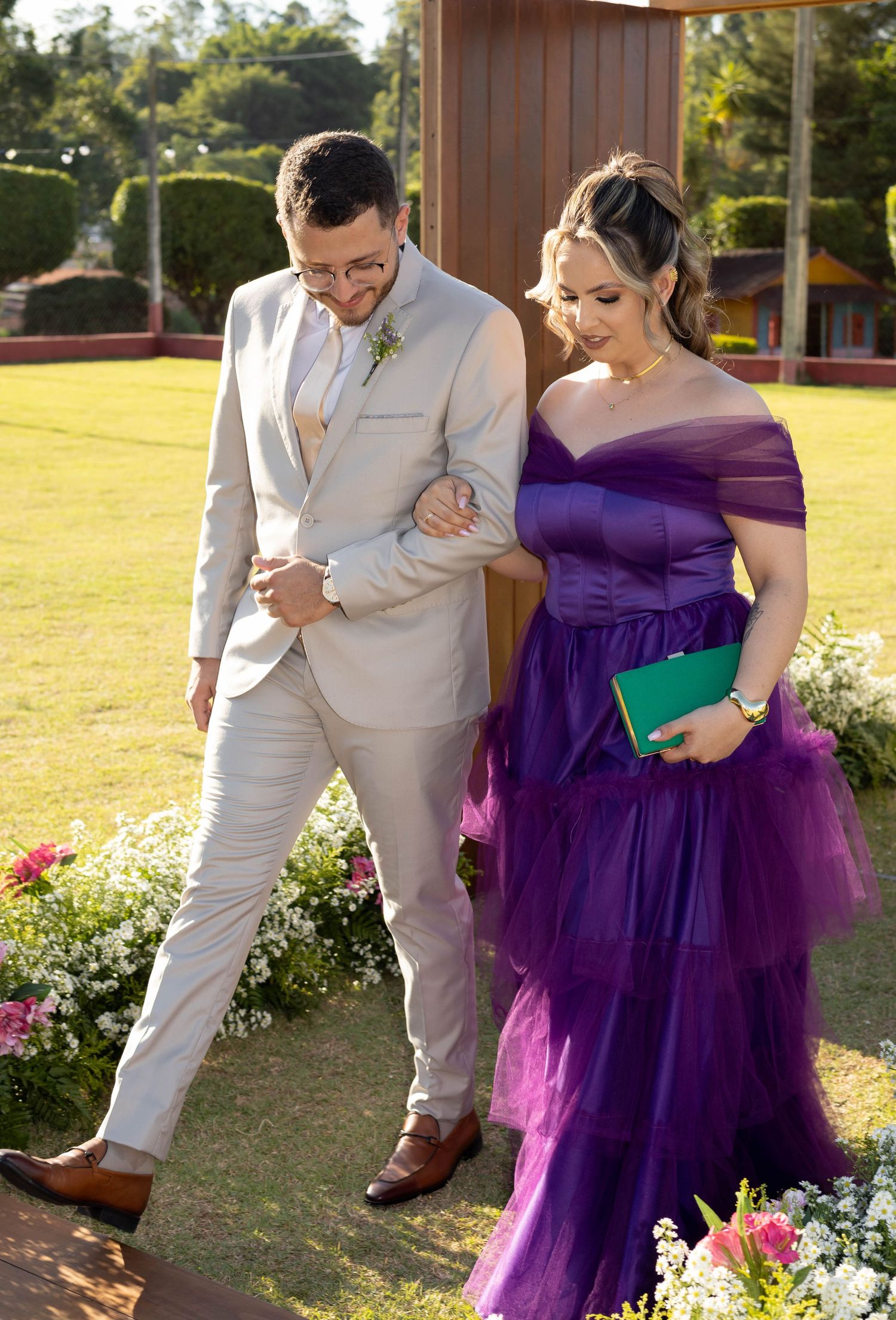 Couple in formal attire, man in beige suit and woman in purple off-shoulder gown, walking at outdoor wedding.