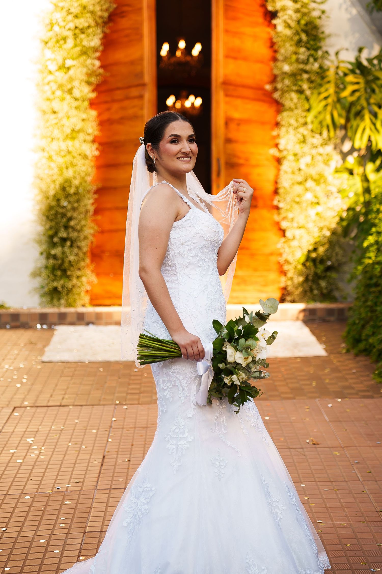 Bride in white mermaid lace gown holding floral bouquet, smiling outdoors near decorated church entrance.
