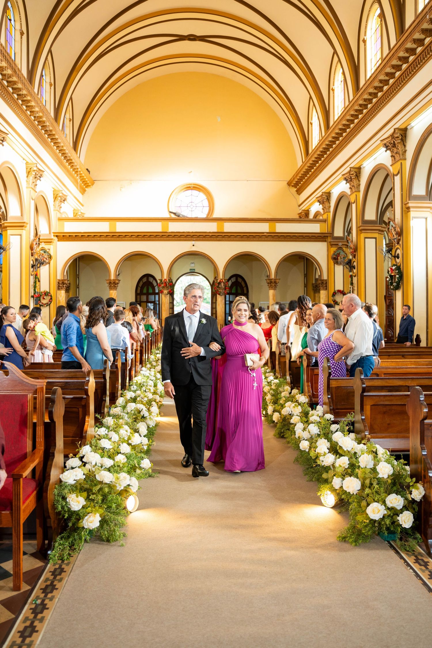 Couple walks down floral-lined church aisle, woman in magenta gown, man in suit, guests seated in pews.
