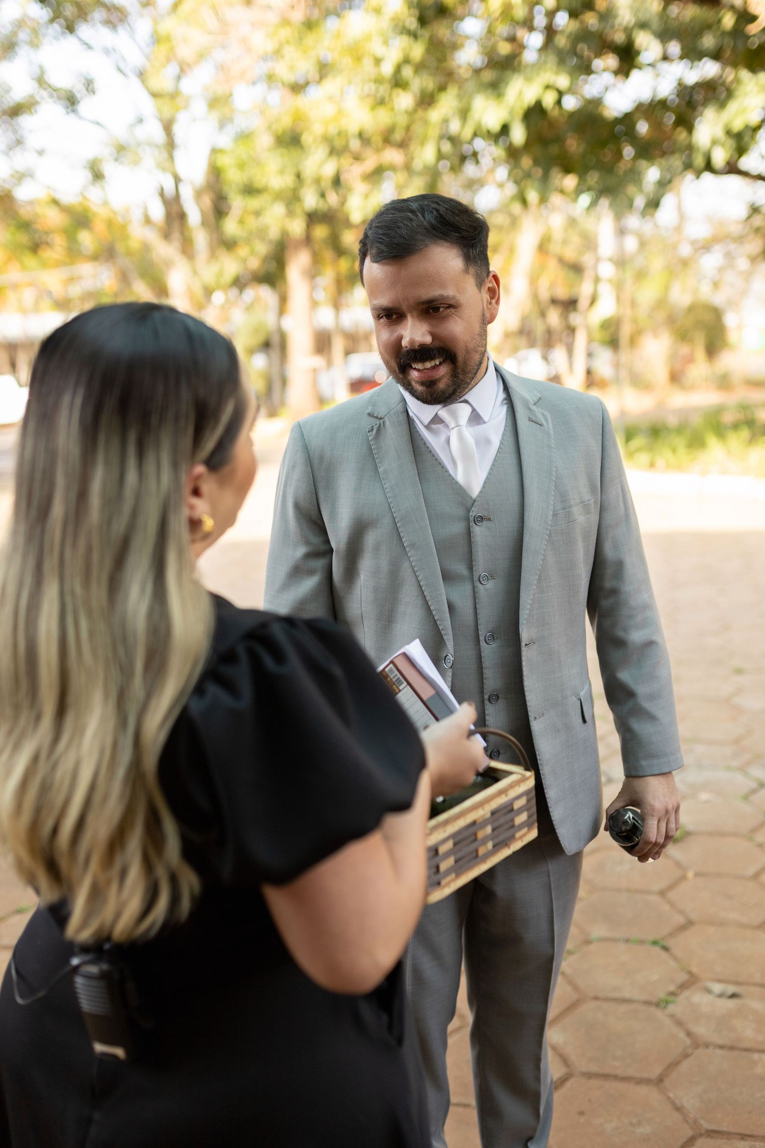 Man in gray suit smiling at blonde woman in black dress outdoors, both holding books in a sunny park setting.
