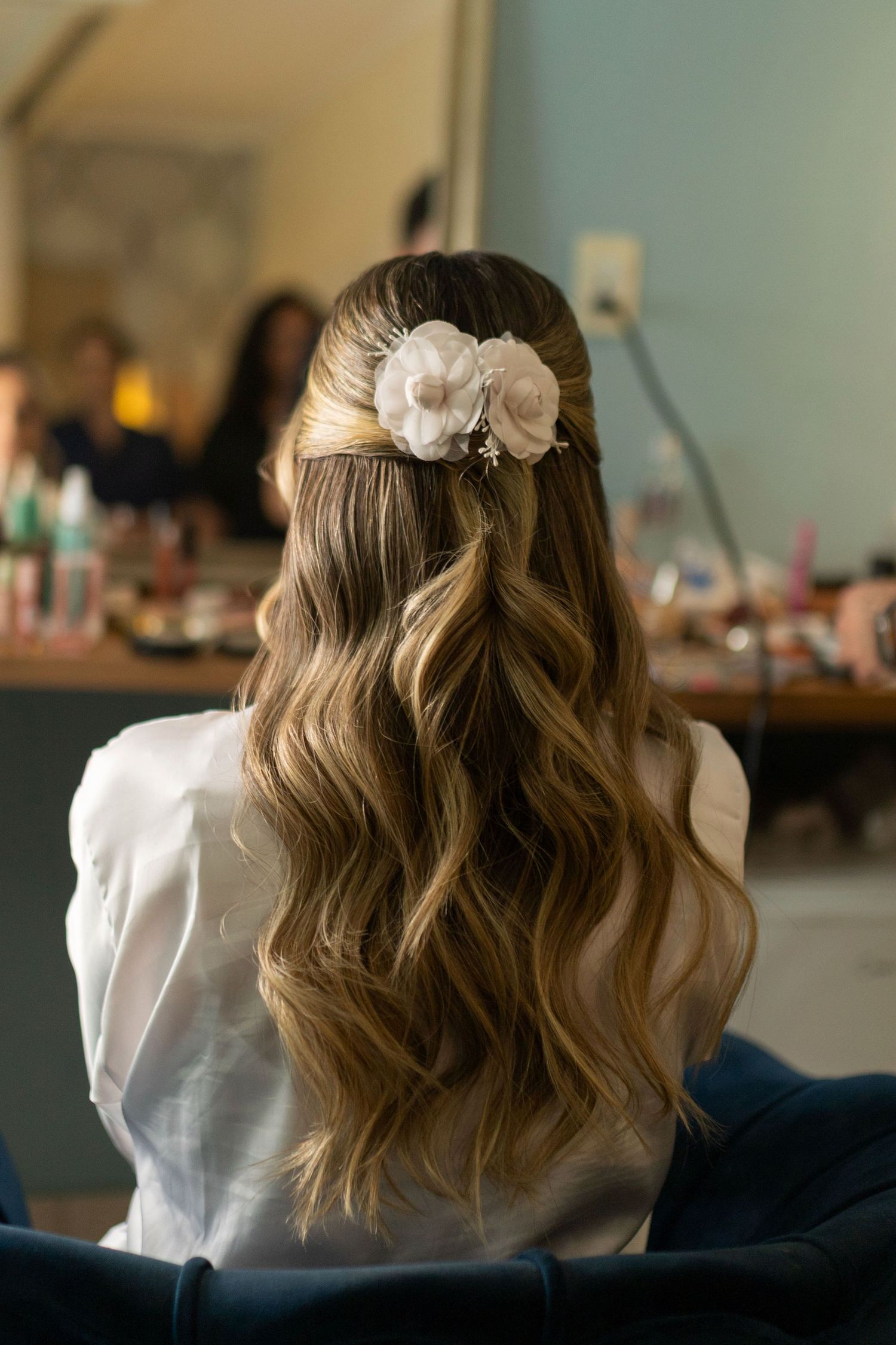 Bride with long wavy hair adorned with white floral hair clip, half-up style, sitting in bridal suite.