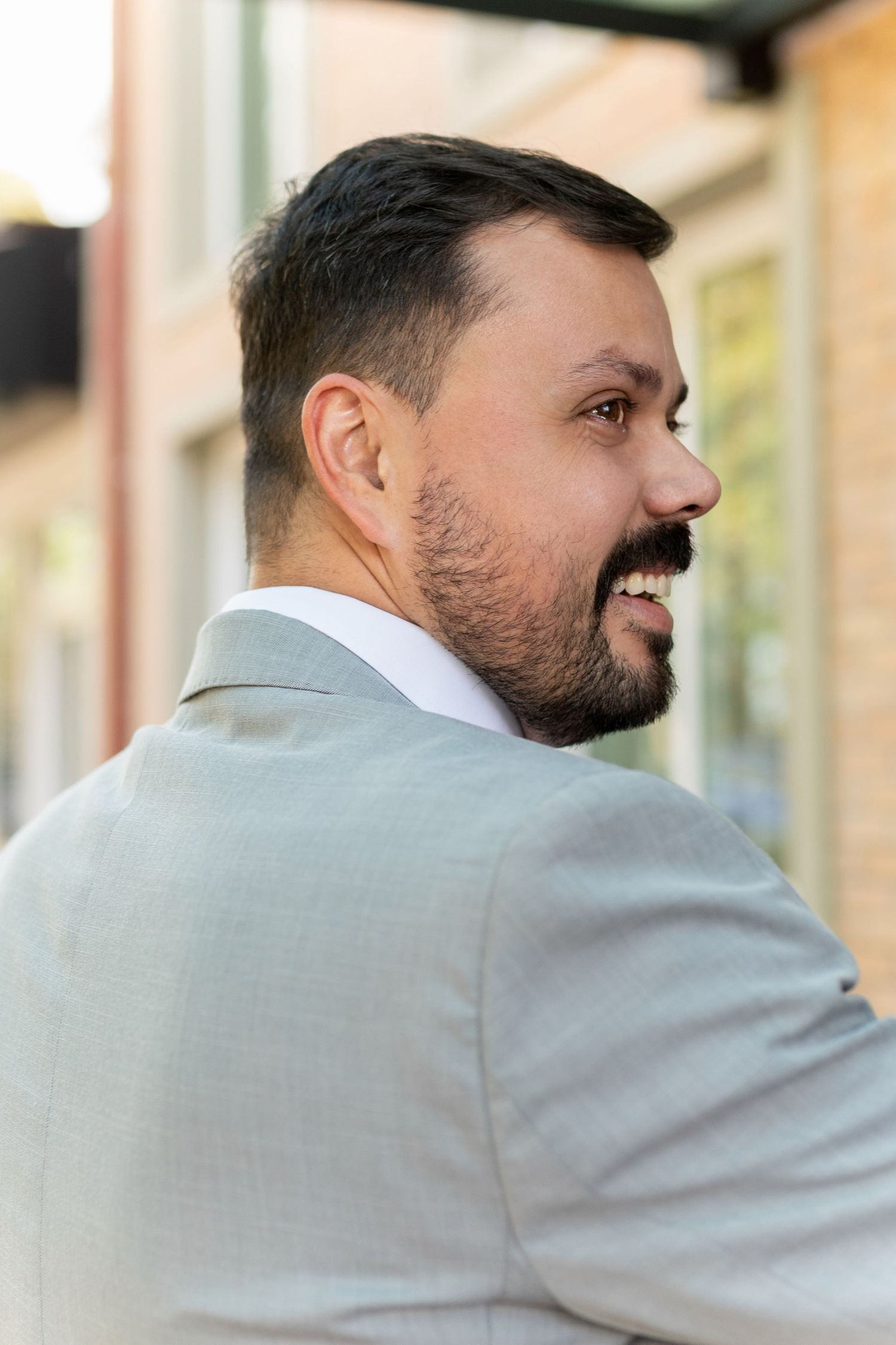 Smiling bearded man in a light gray suit photographed from behind, turning to show his profile outdoors.