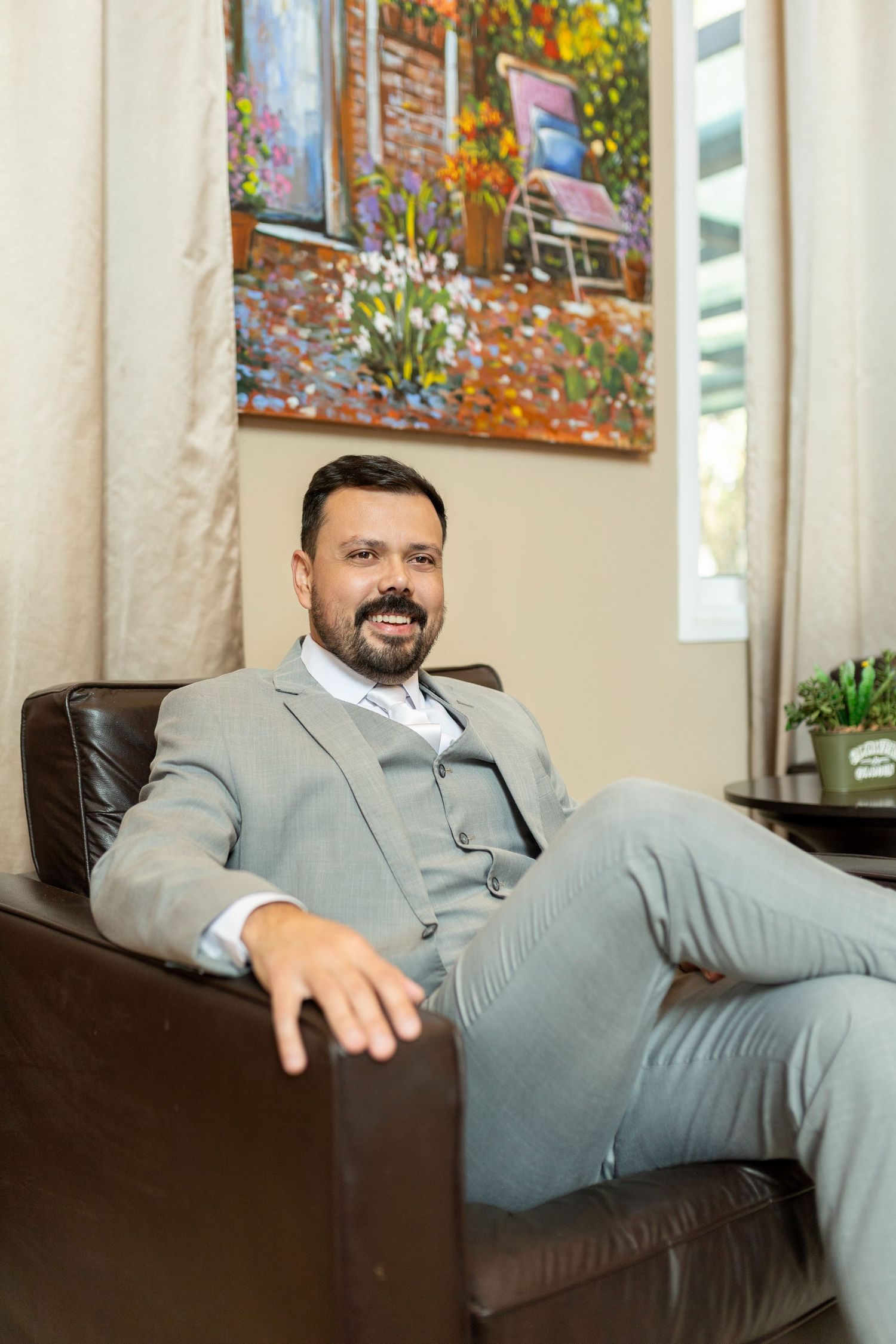 A smiling bearded man in a gray suit sits relaxed in a leather chair before a colorful floral painting.