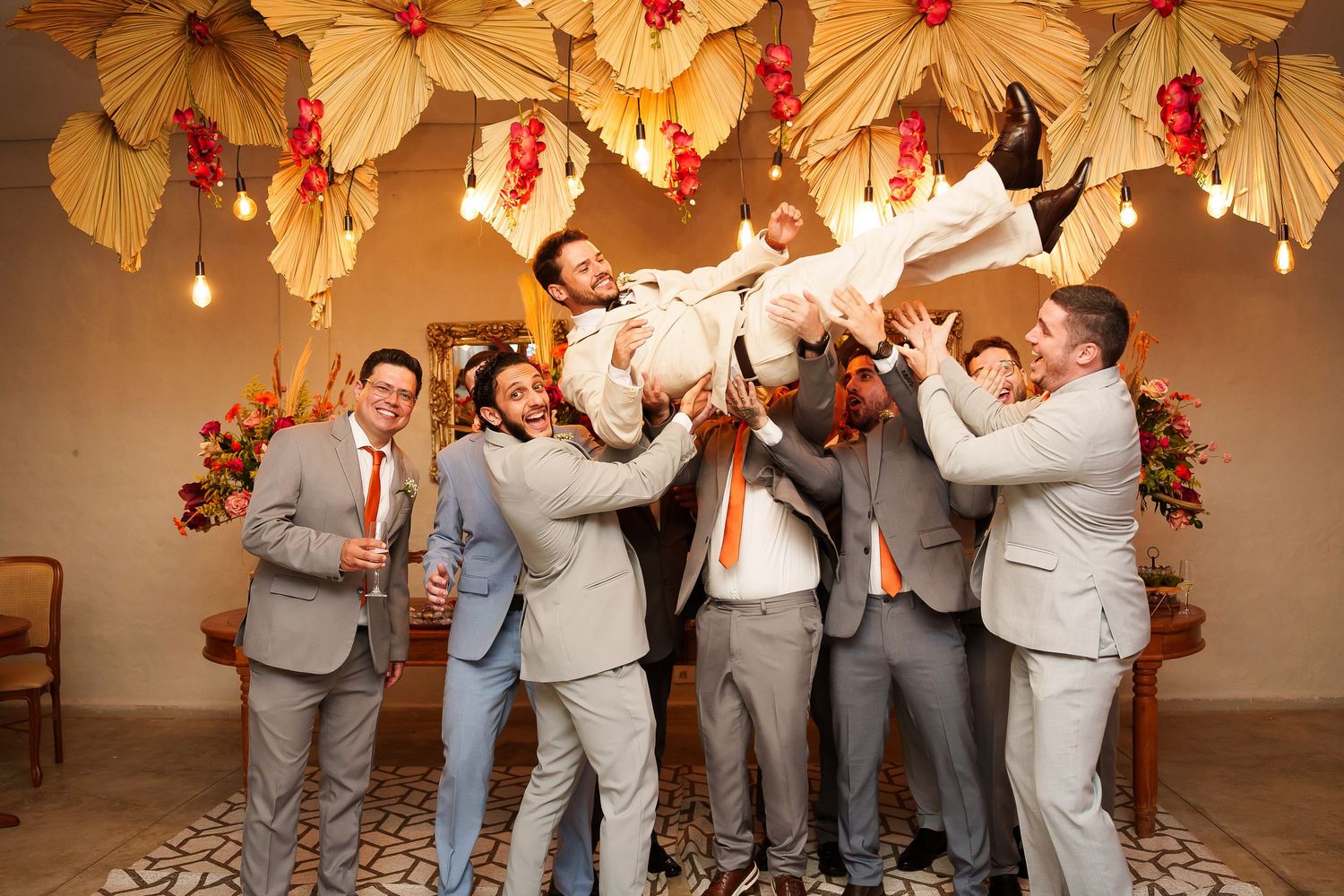 Groomsmen in gray suits lifting groom in white suit at decorated wedding venue with floral ceiling decor.
