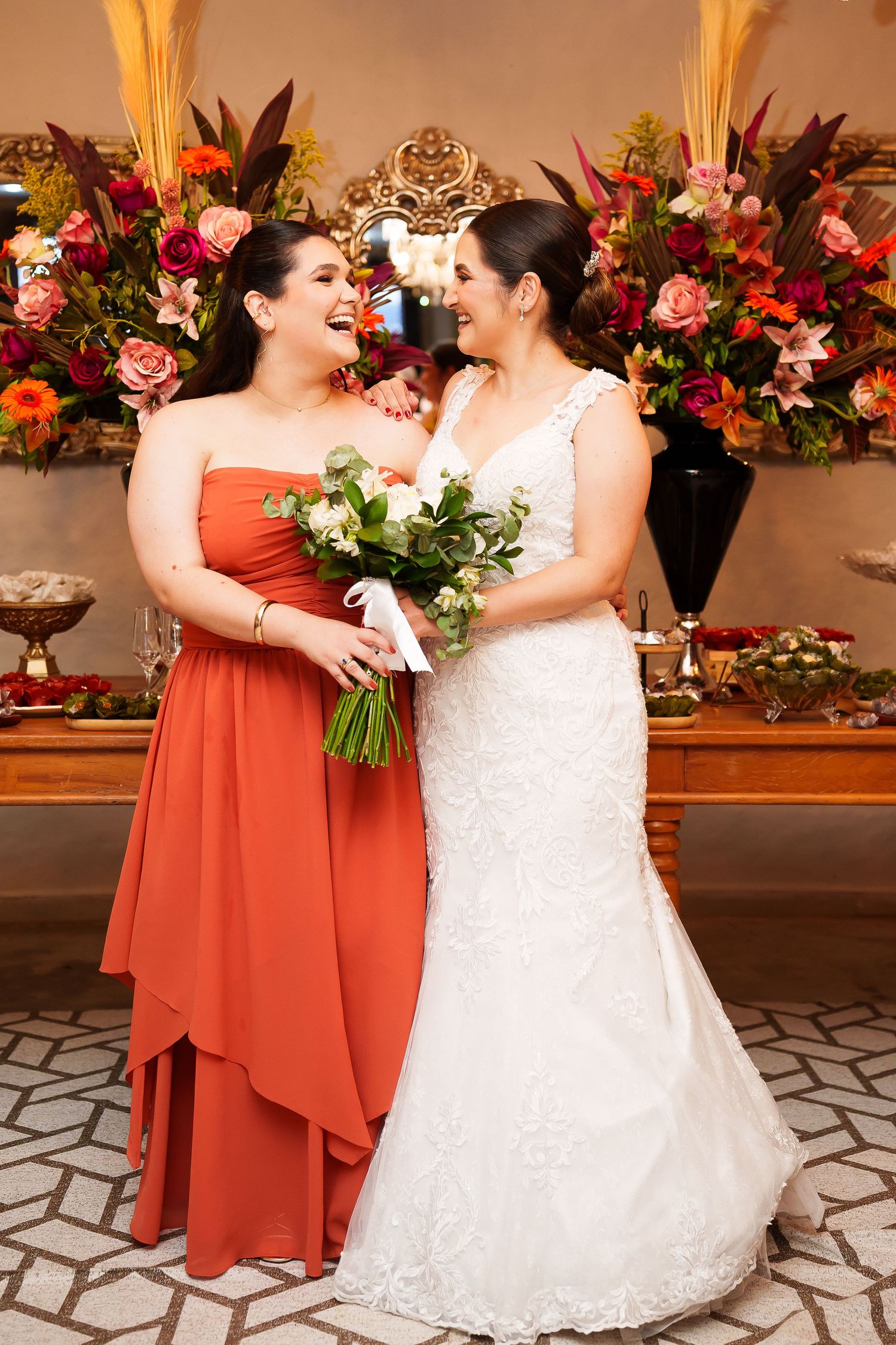 Bride in white lace gown and bridesmaid in orange dress laughing together, holding bouquet at wedding reception.