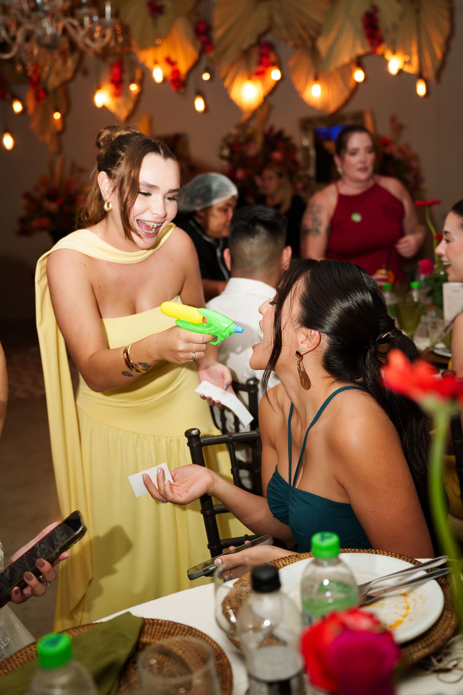 Woman in yellow dress playfully uses water gun at festive party, guests laughing at decorated venue.