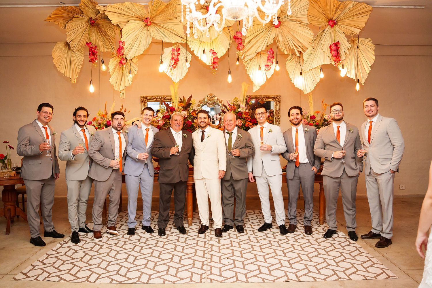 Groom in white suit poses with groomsmen in gray suits and orange ties under floral decor at elegant wedding venue.