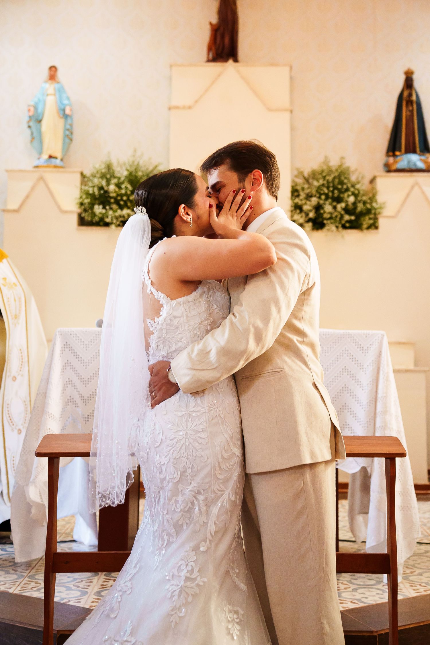 Bride and groom share first kiss at church altar, bride in lace gown with veil, groom in beige suit.