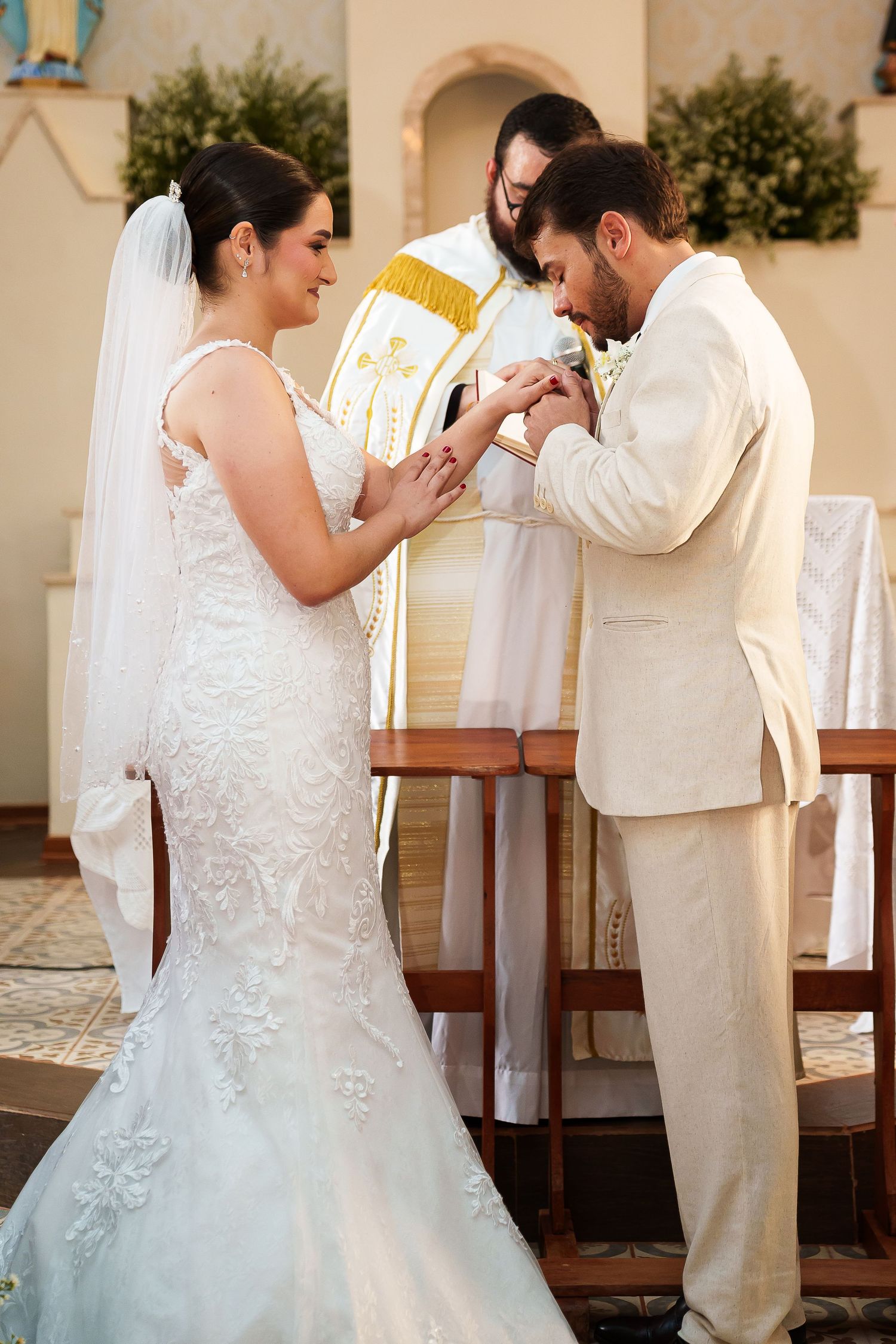 Bride and groom exchange rings at church altar during wedding ceremony, officiated by priest in gold vestments.
