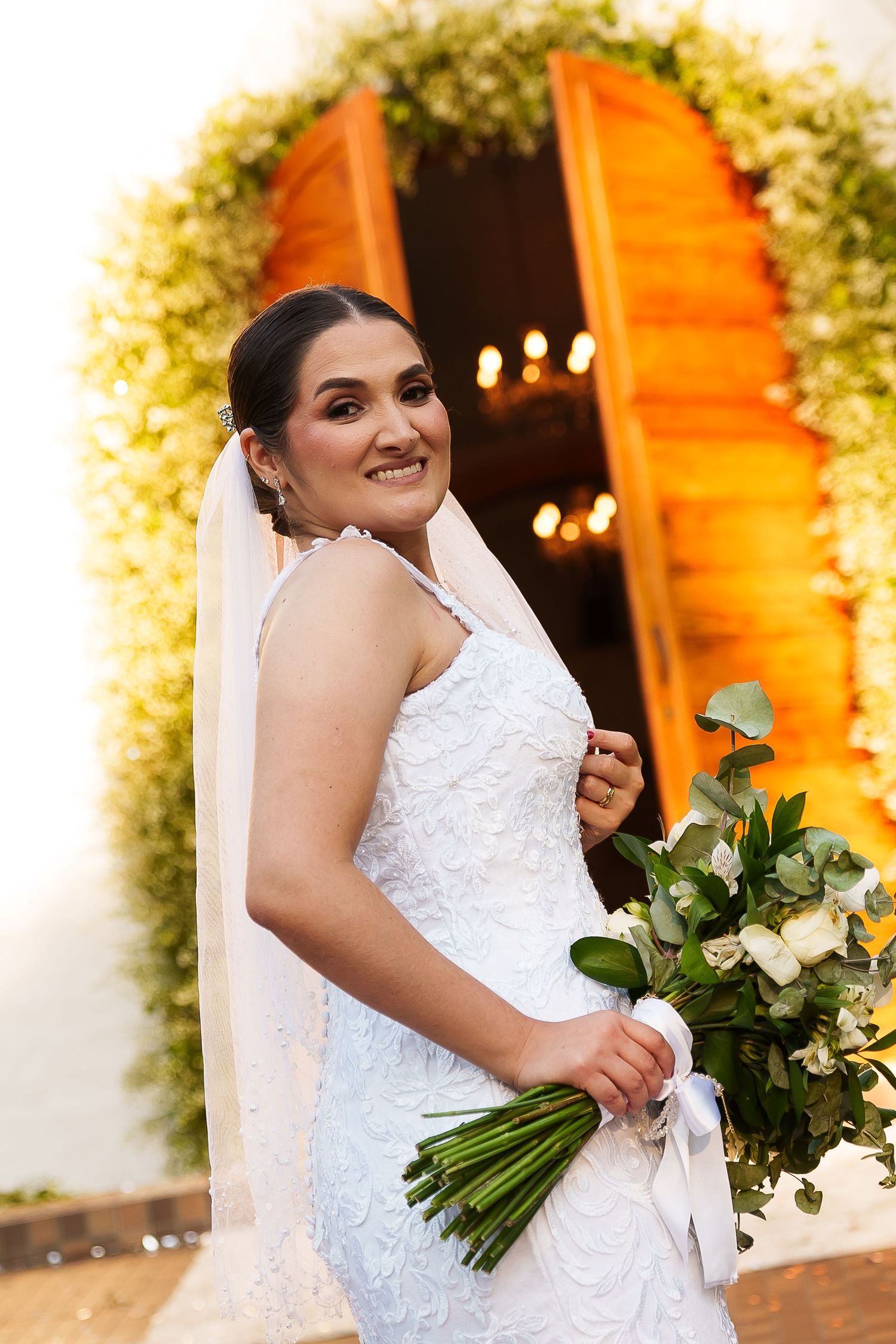 Smiling bride in white lace gown and veil holds bouquet before floral arch with warm lighting.