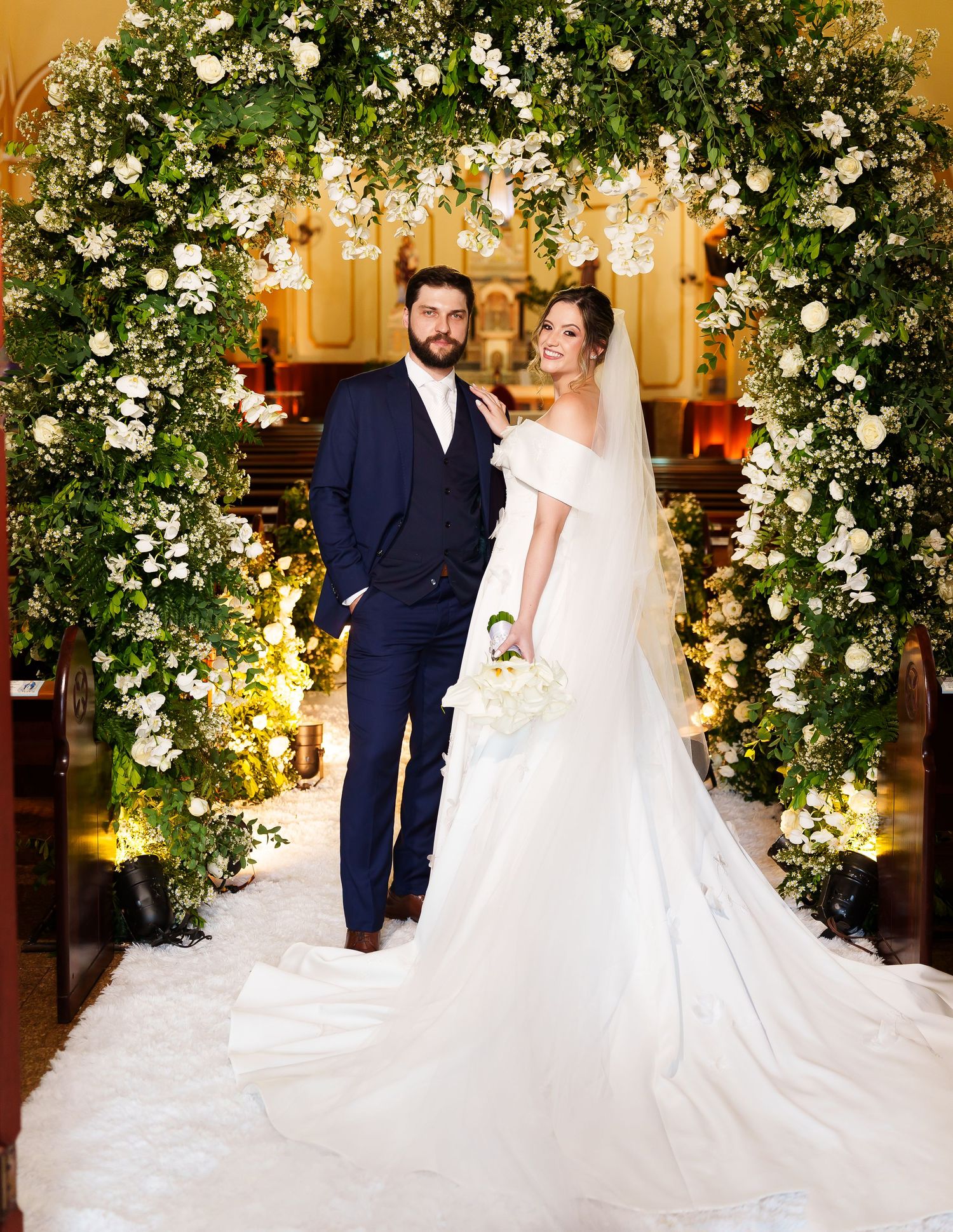 Bride and groom posing under a floral arch of white roses and greenery inside a church on their wedding day.