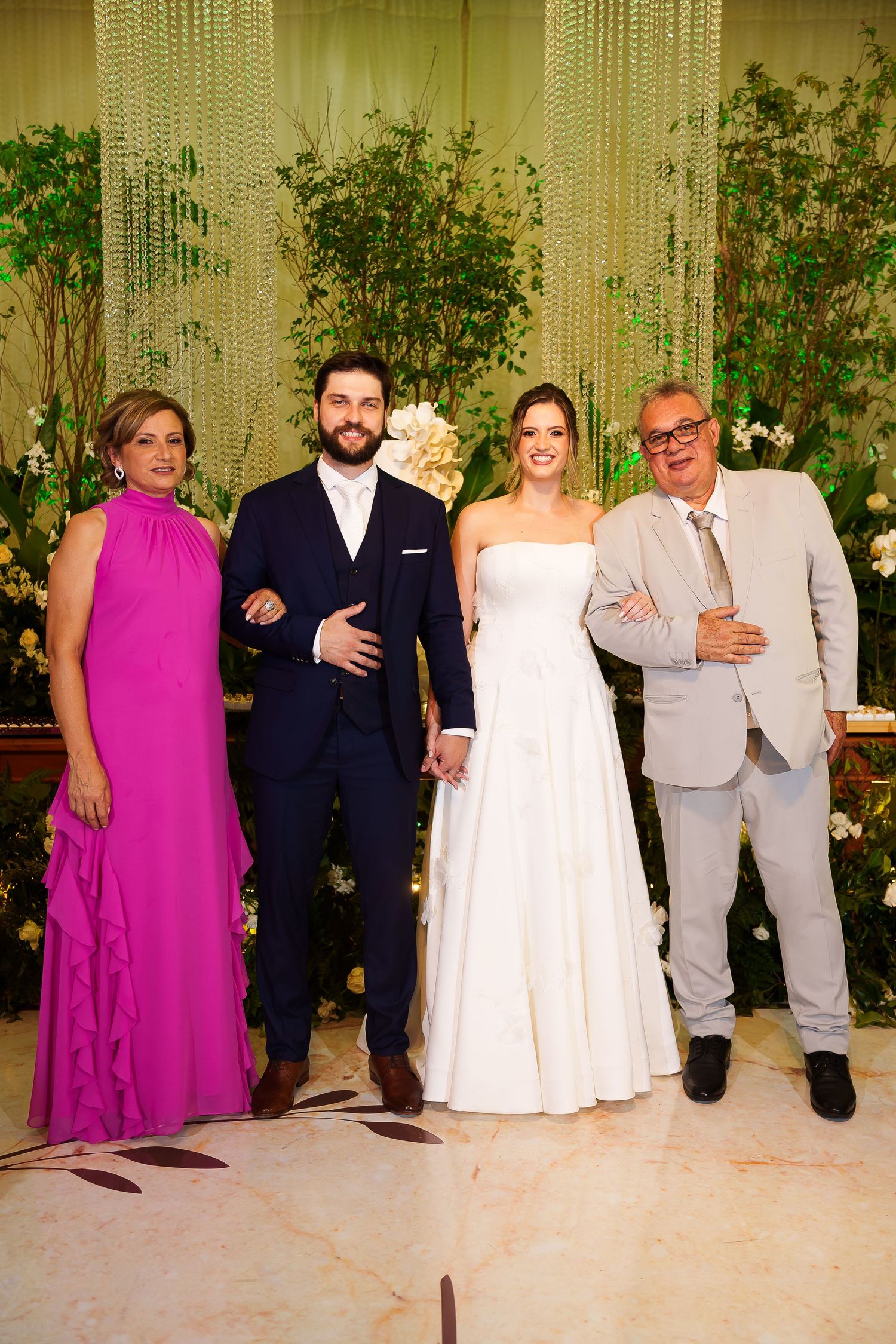 Wedding couple poses with family, bride in white gown, groom in navy suit, woman in pink dress, man in gray suit.