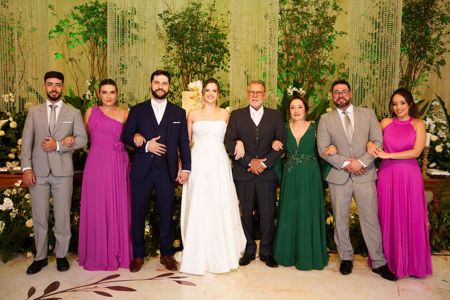 Wedding party poses together in elegant venue, bride in white gown flanked by family in purple, green, and gray formal attire.