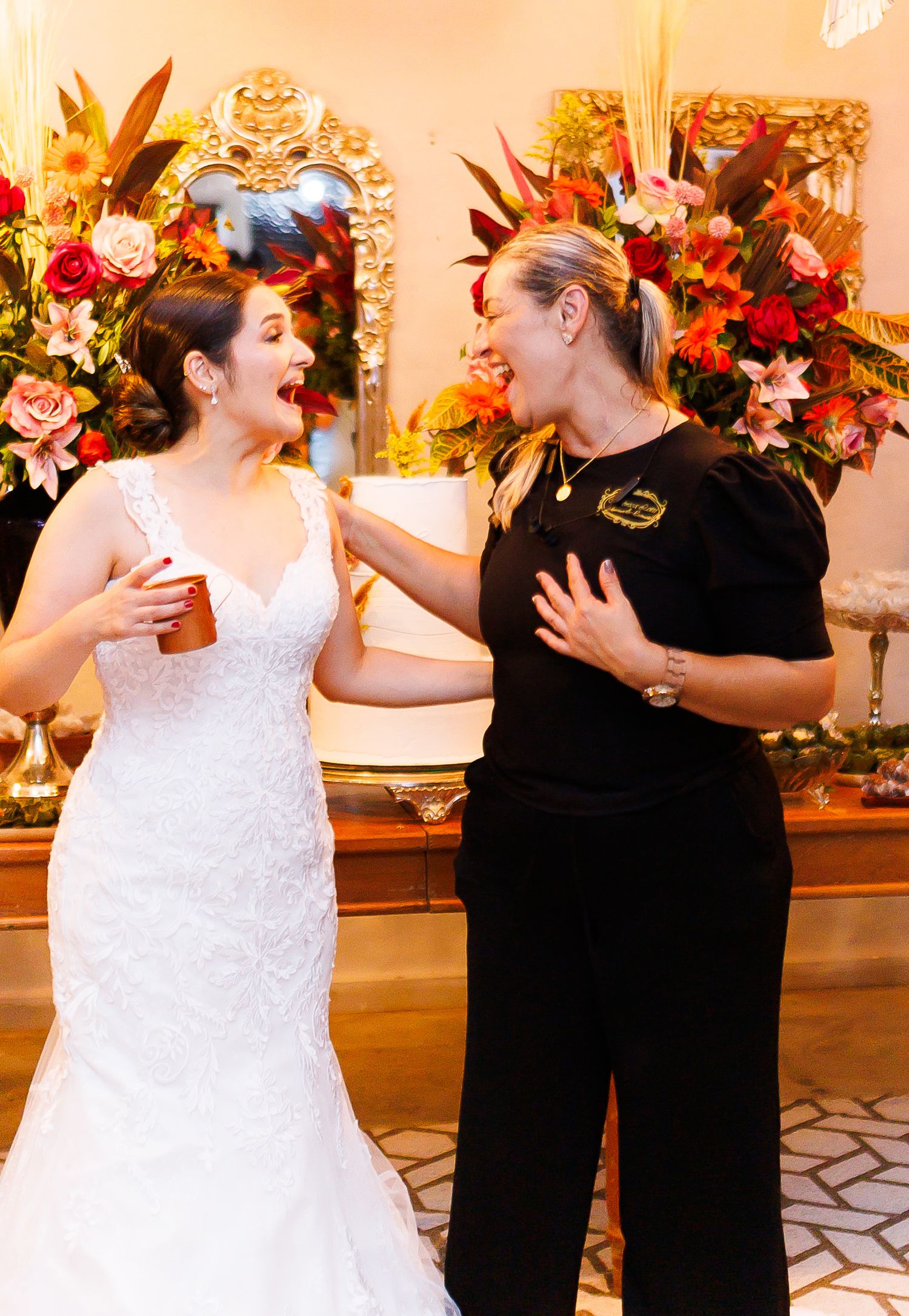 Bride in white lace gown laughing with woman in black top at wedding reception with floral backdrop.
