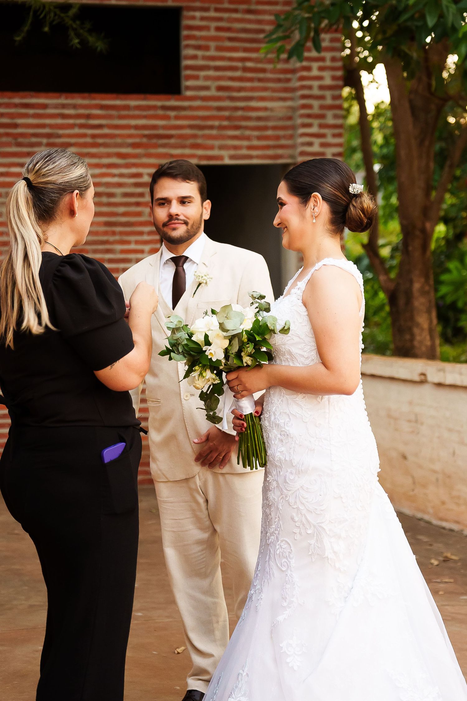 Bride in white gown holding bouquet speaks with officiant as groom in beige suit stands outdoors near brick wall.