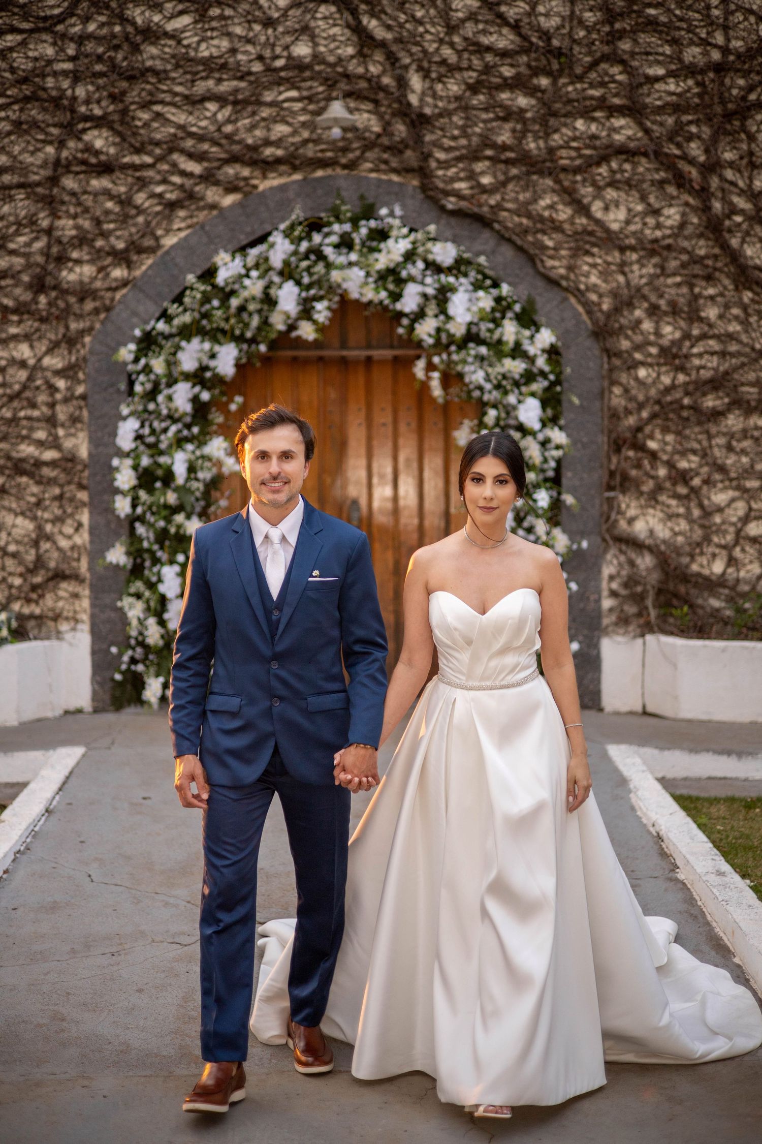 Bride and groom holding hands before a floral arch doorway, bride in strapless gown, groom in navy suit.