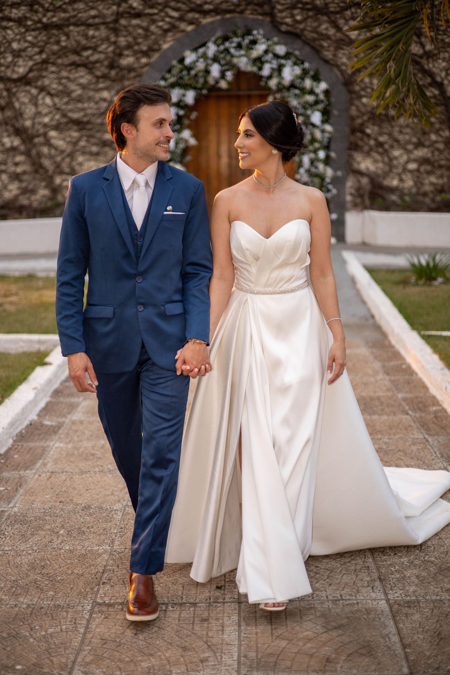 A bride in a white strapless gown and groom in a blue suit hold hands walking toward a floral arch.