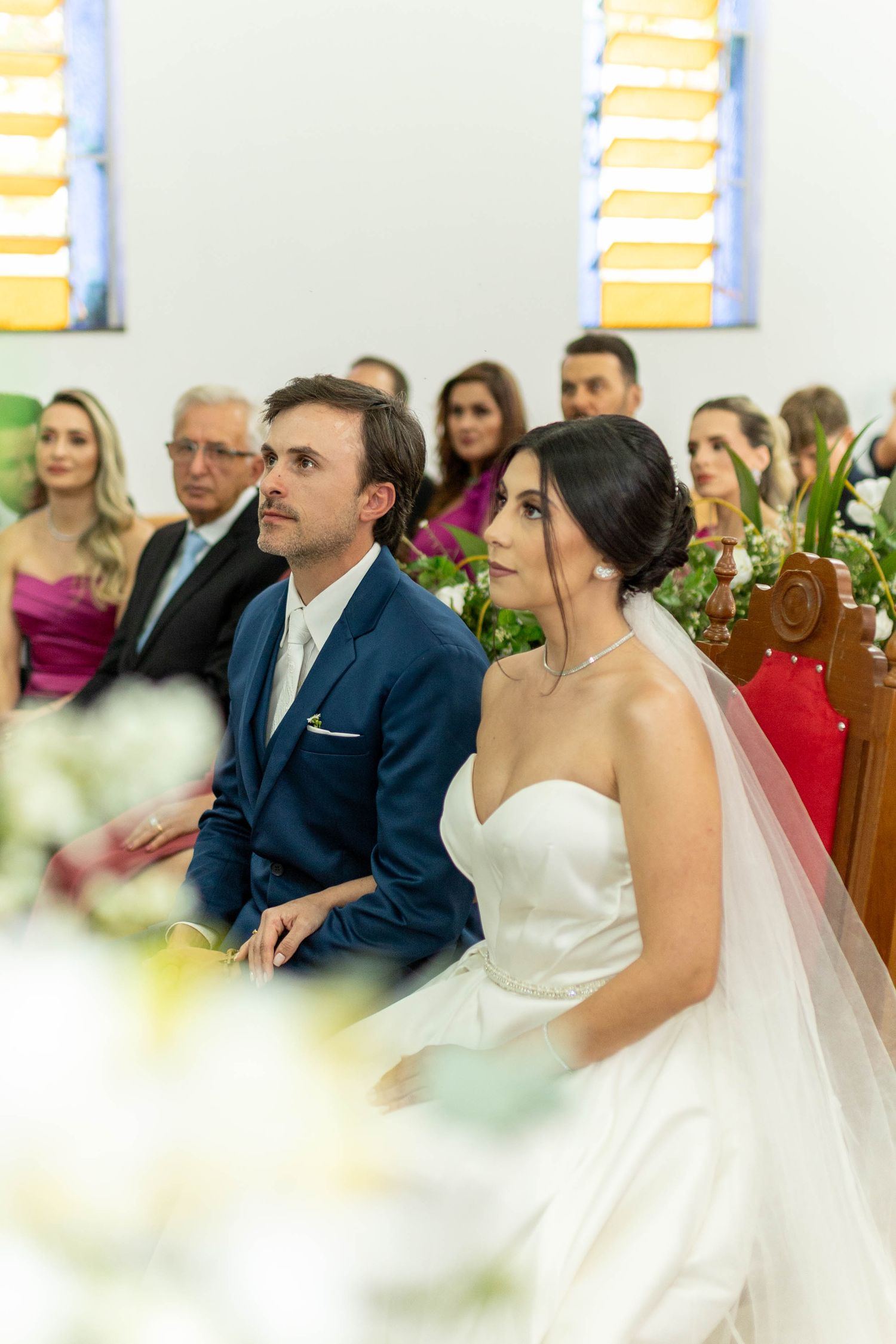 Bride in white gown and groom in blue suit sitting at church altar during wedding ceremony with guests in background.