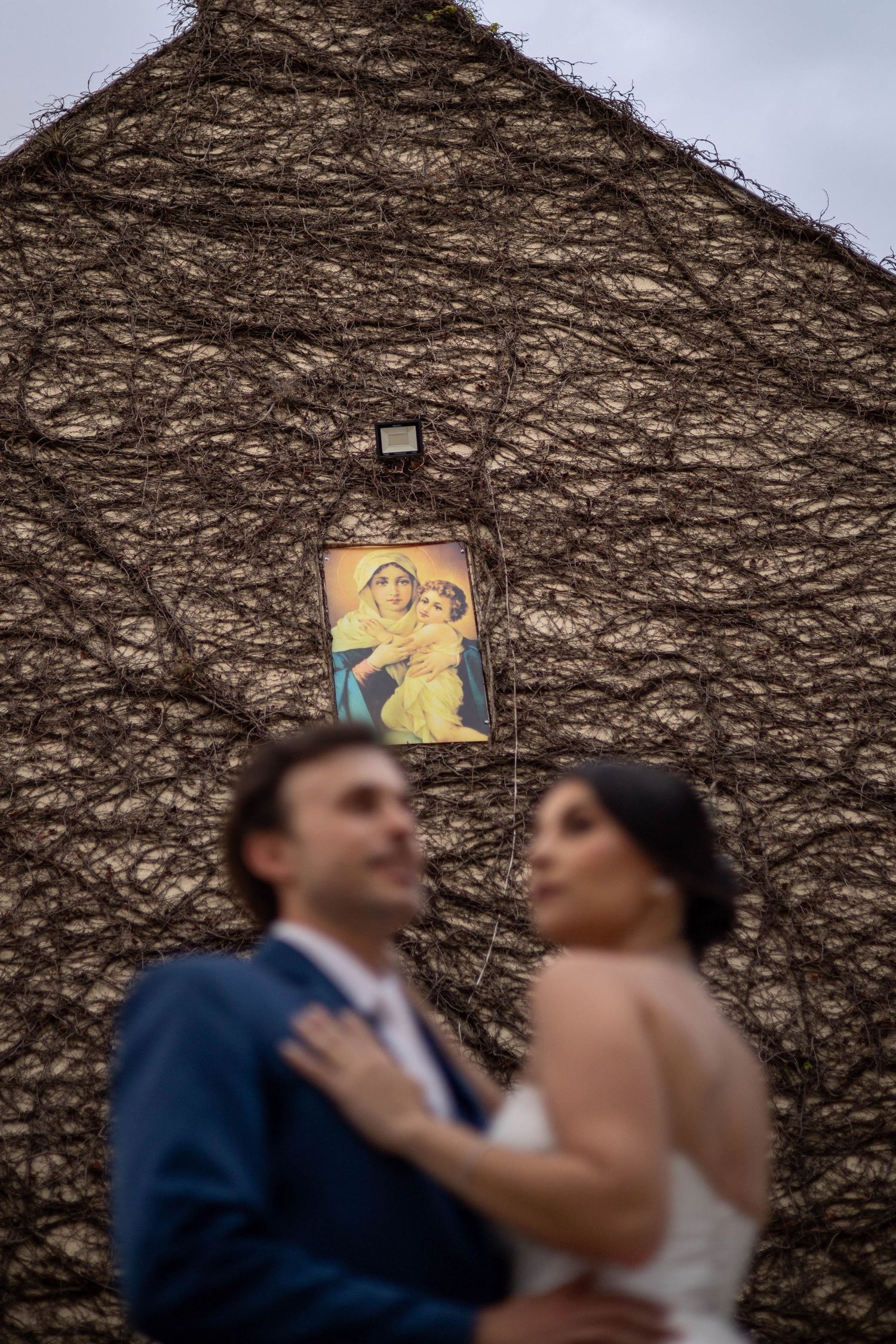 Blurred bride and groom pose romantically before a vine-covered wall with a Virgin Mary icon above them.