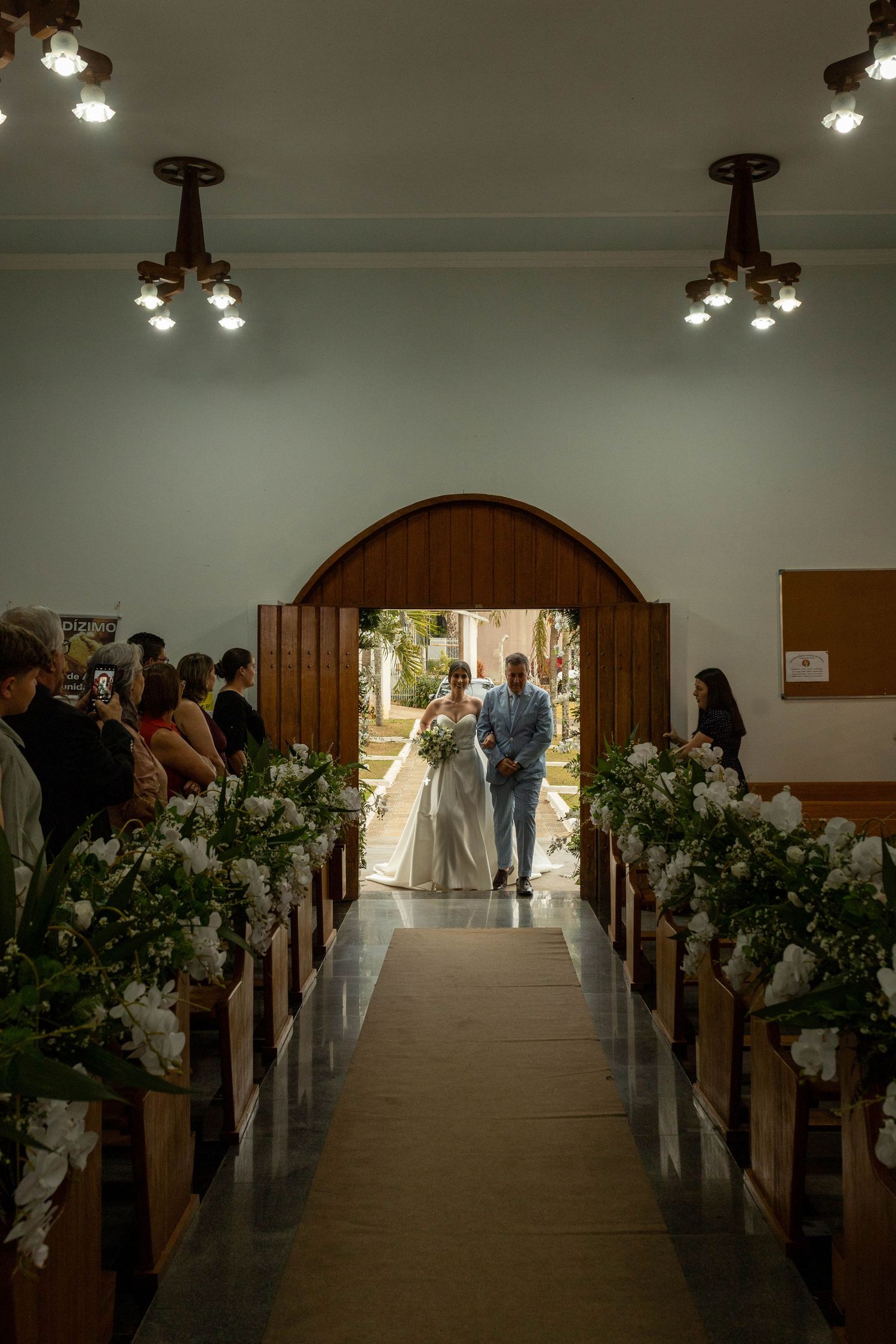 Bride walks down the aisle with escort in a decorated church with floral arrangements on wooden pews.