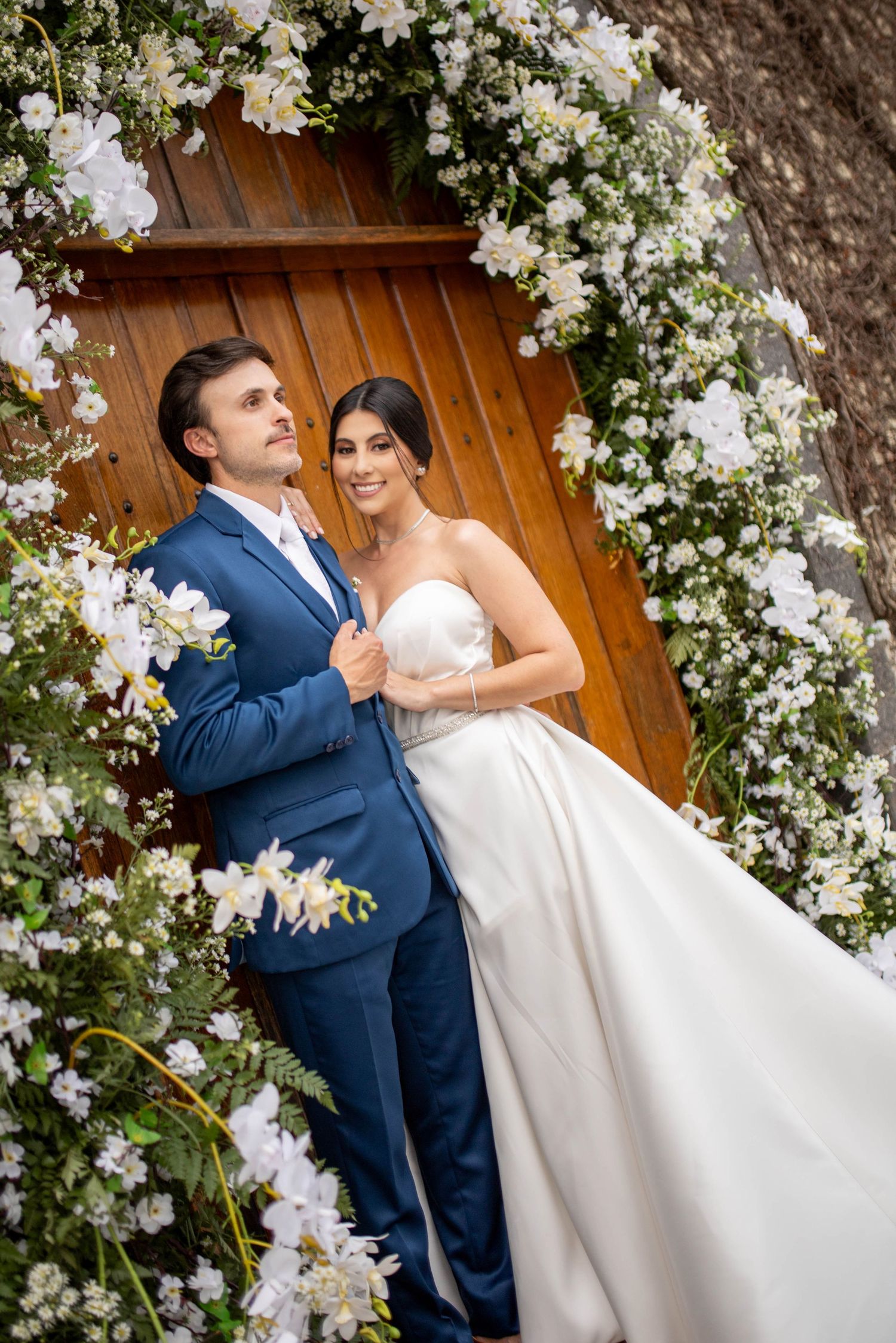 Bride in white strapless gown and groom in blue suit pose before floral arch with white flowers and wooden door.