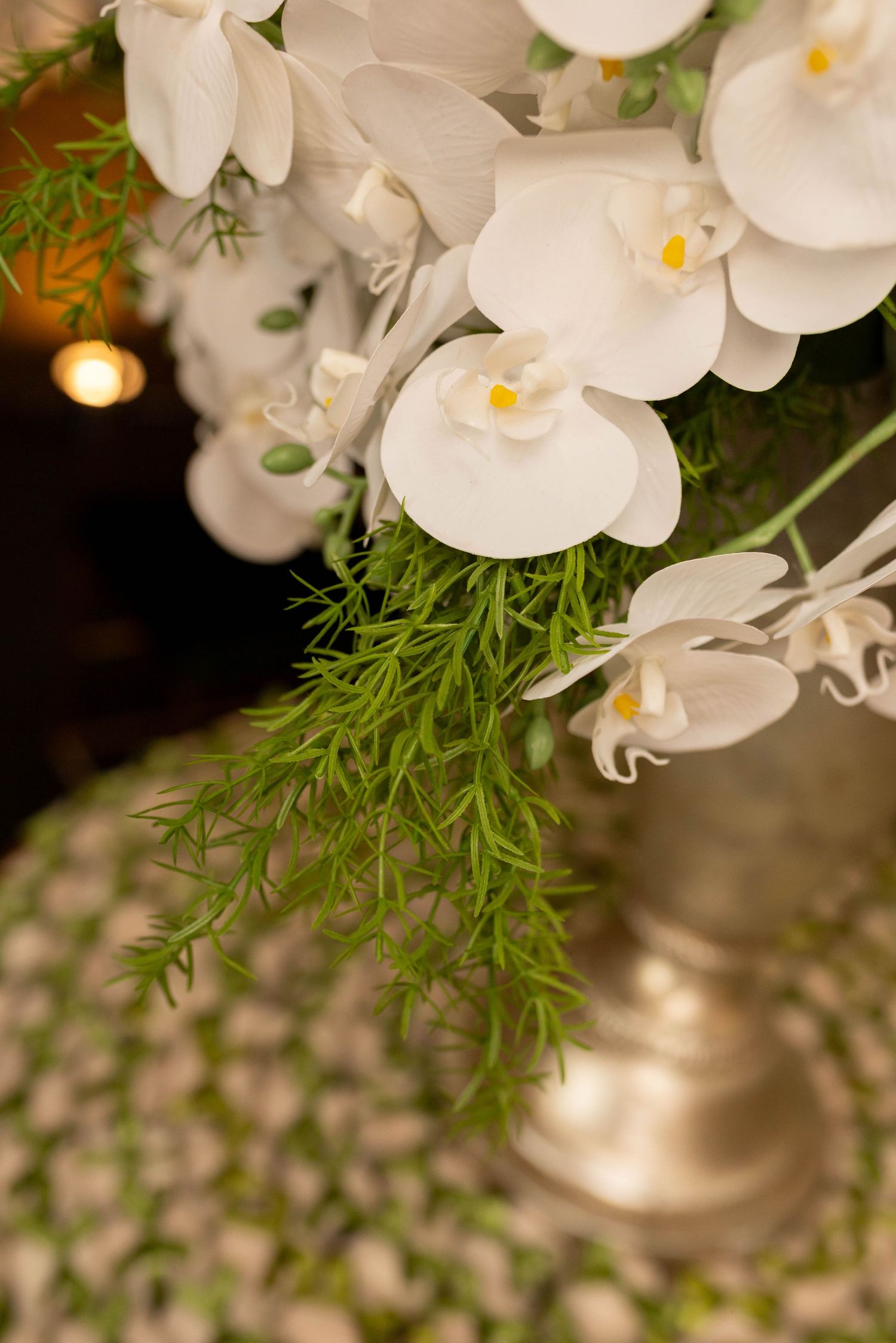 White phalaenopsis orchids arranged with green ferns in a silver vase, elegant floral centerpiece.
