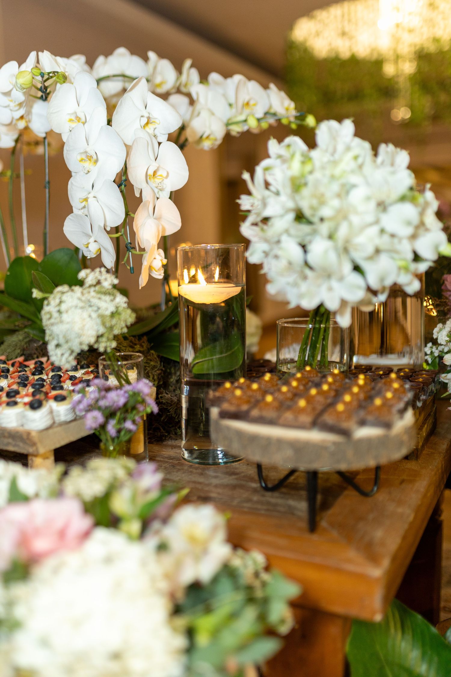 Elegant wedding dessert table with white orchids, candles, and chocolate treats on rustic wood stands.