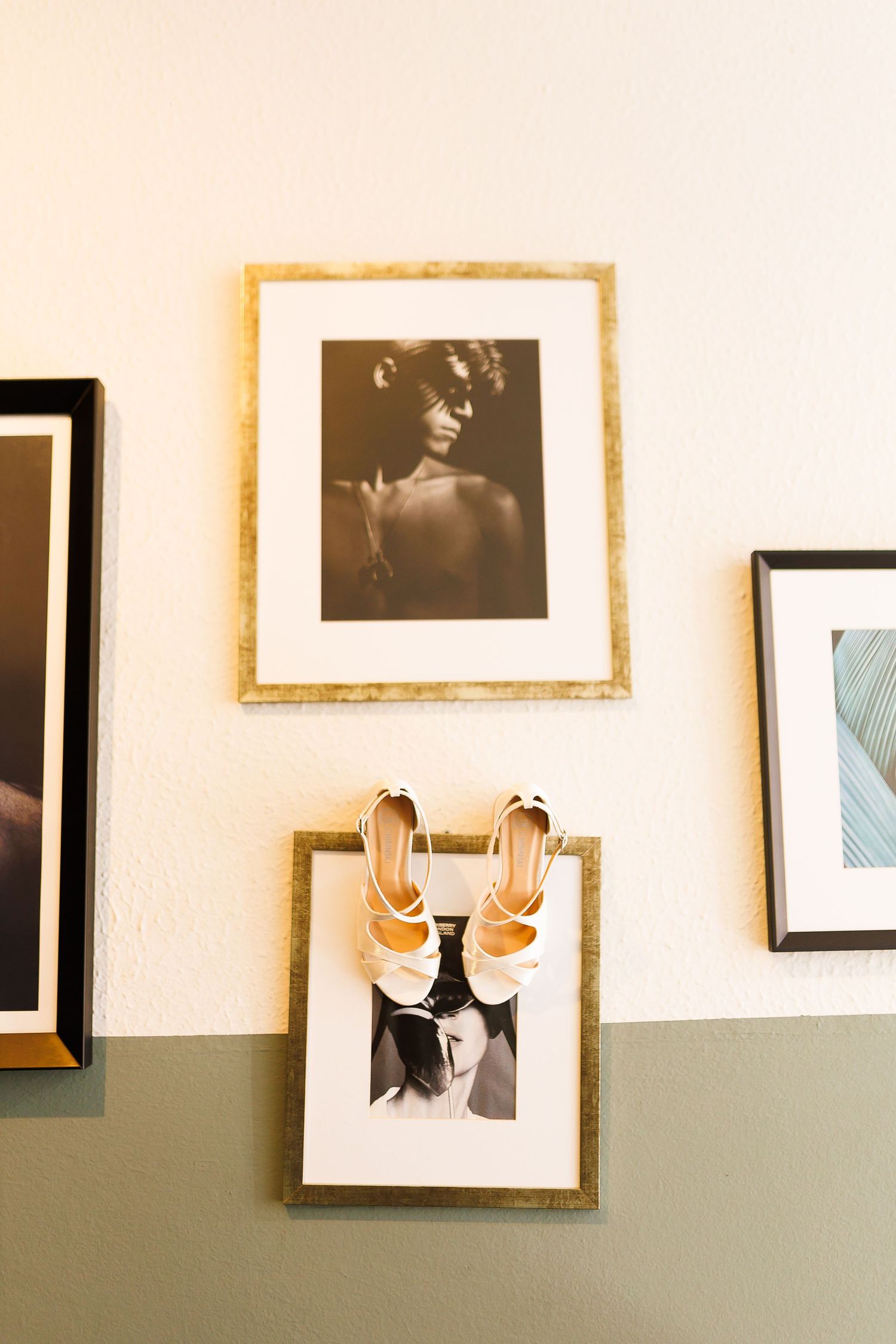 White heeled sandals displayed on gold-framed black and white portrait photos hanging on a two-toned wall.