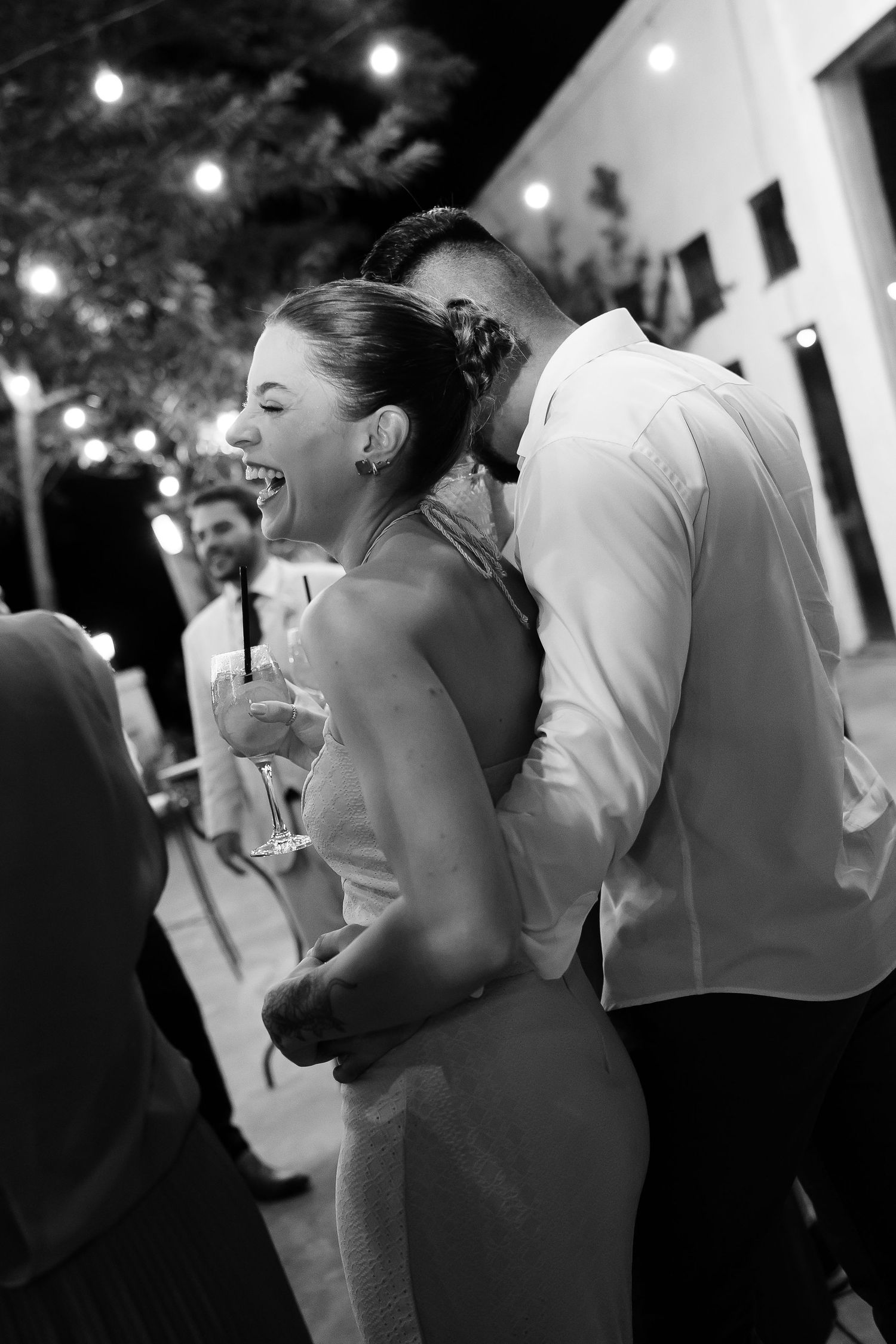 Black and white photo of a laughing bride holding champagne while groom whispers in her ear at outdoor wedding reception.
