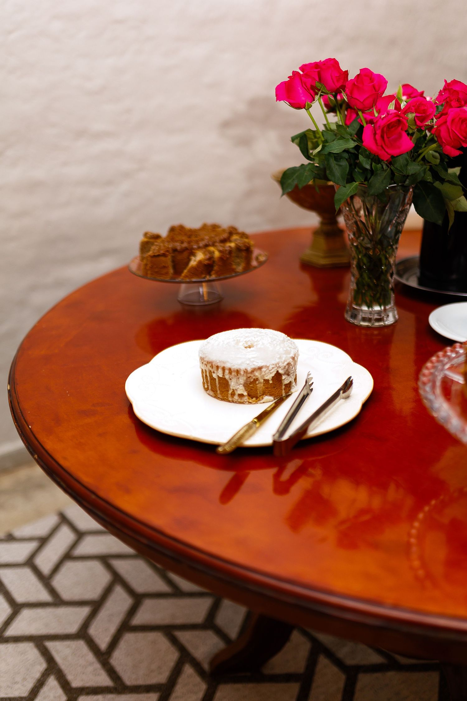 Elegant round wooden table with a frosted pastry, gold cutlery, pink roses bouquet, and cake on display stand.