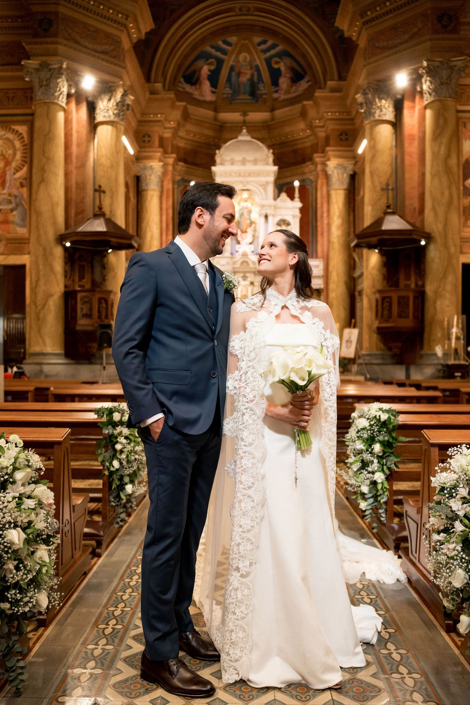 Bride and groom share a romantic moment at church altar, she holds white bouquet in elegant lace wedding dress.