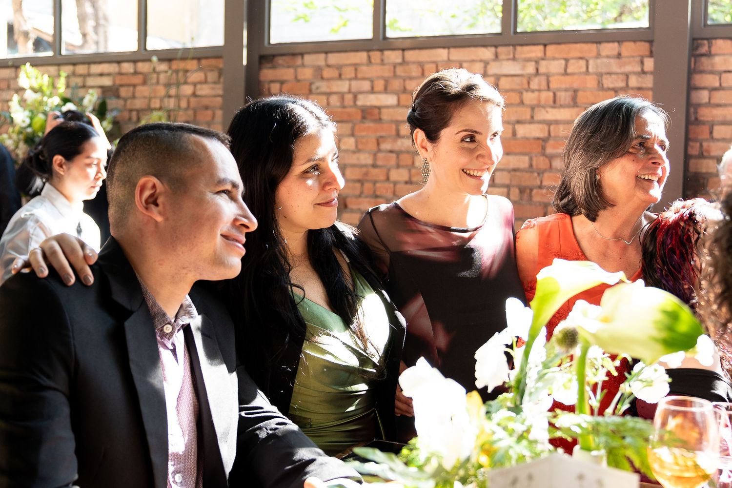 Group of smiling guests seated at a table during an indoor wedding reception near a brick wall.