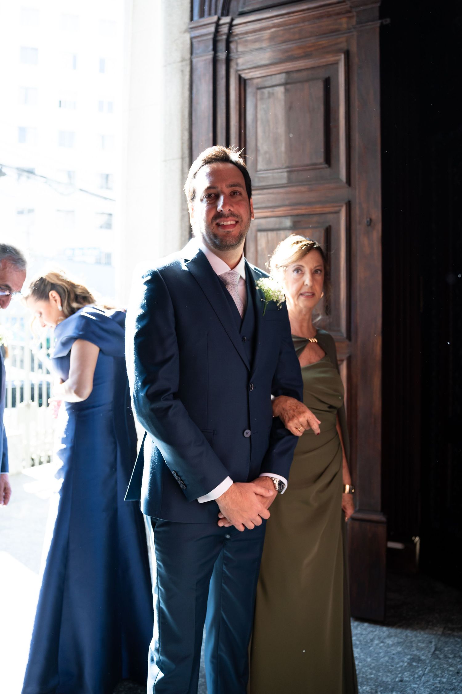 Groom in navy suit stands with woman in olive green gown near wooden doors at an elegant wedding venue.