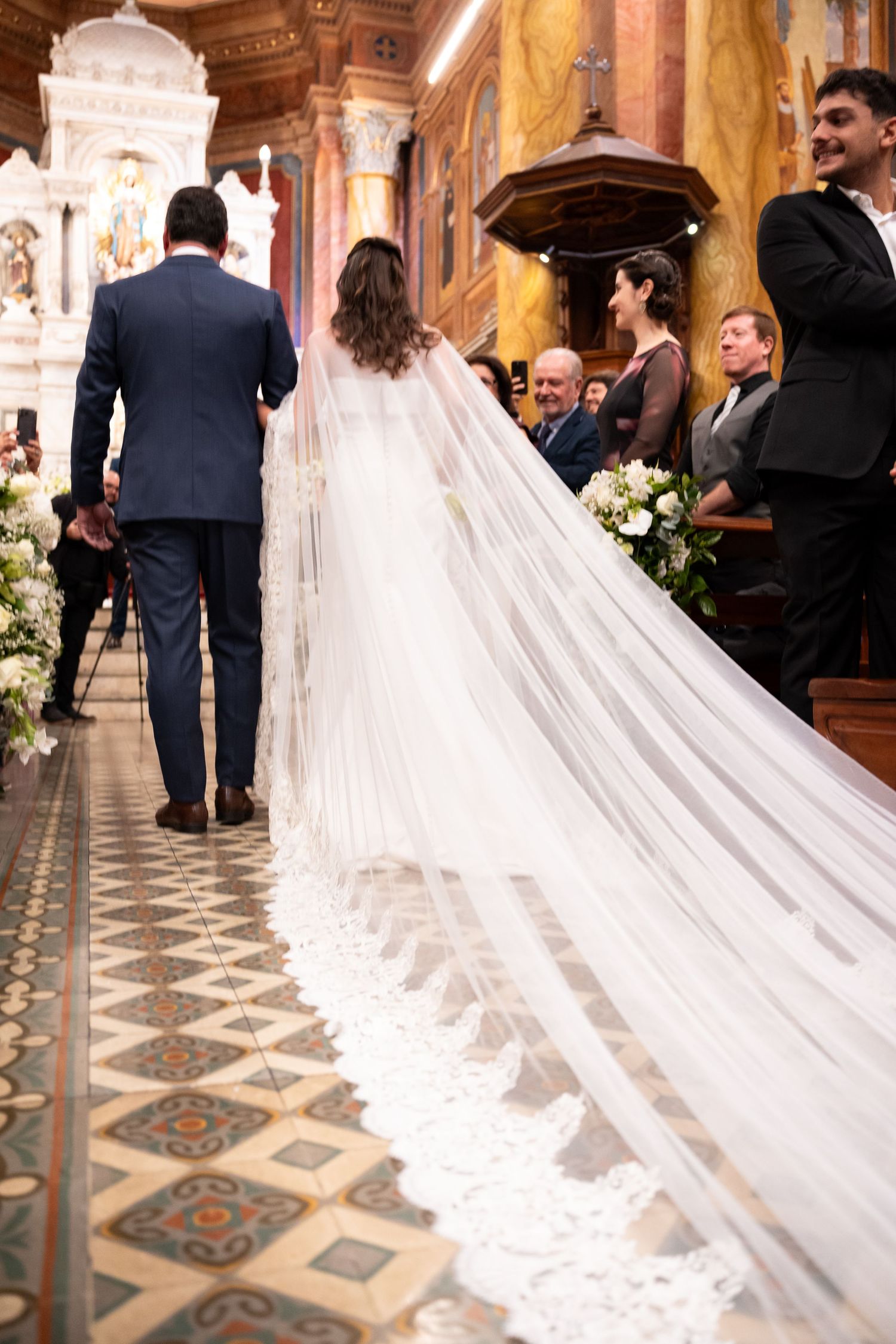 Bride in lace wedding gown with cathedral veil walks down church aisle with groom in navy suit.