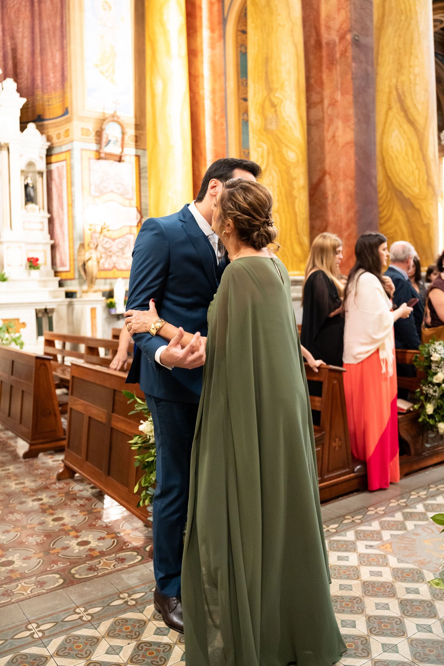 Couple embracing in an elegant church during a wedding ceremony, man in blue suit, woman in green cape dress.