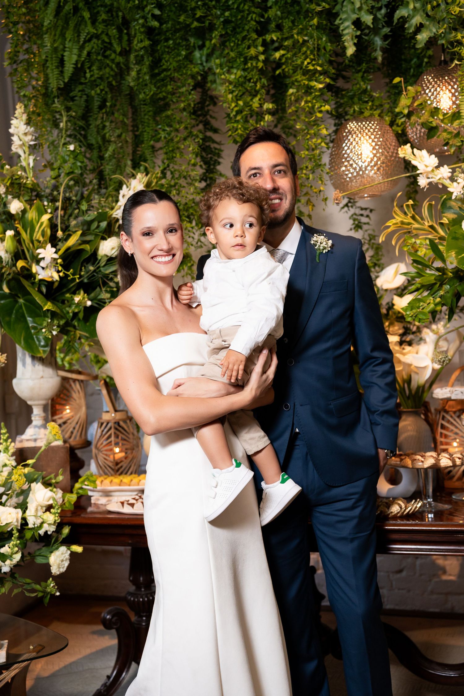 Bride in white gown and groom in navy suit holding young child at elegant wedding reception with greenery backdrop.