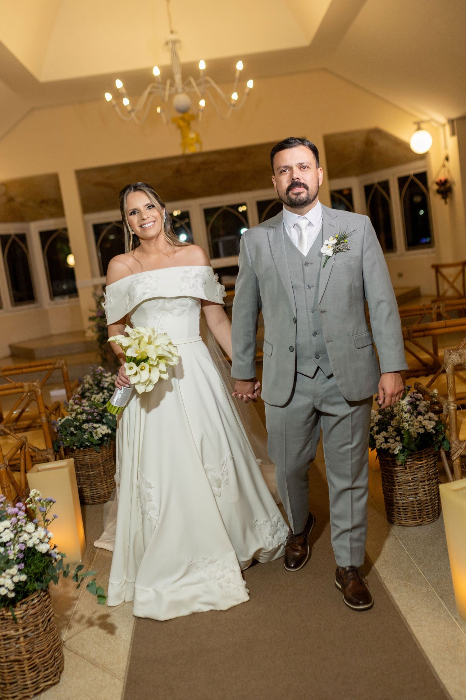 Bride in white off-shoulder gown and groom in gray suit walk down church aisle holding hands with floral decorations.