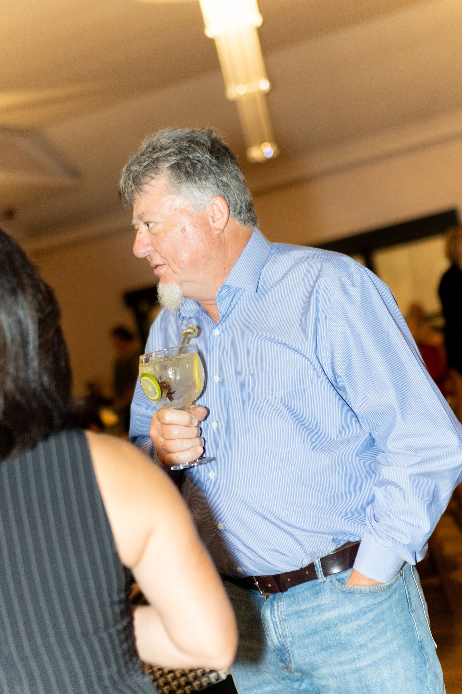 An older man with gray hair holds a cocktail glass at a social gathering wearing a blue shirt and jeans.