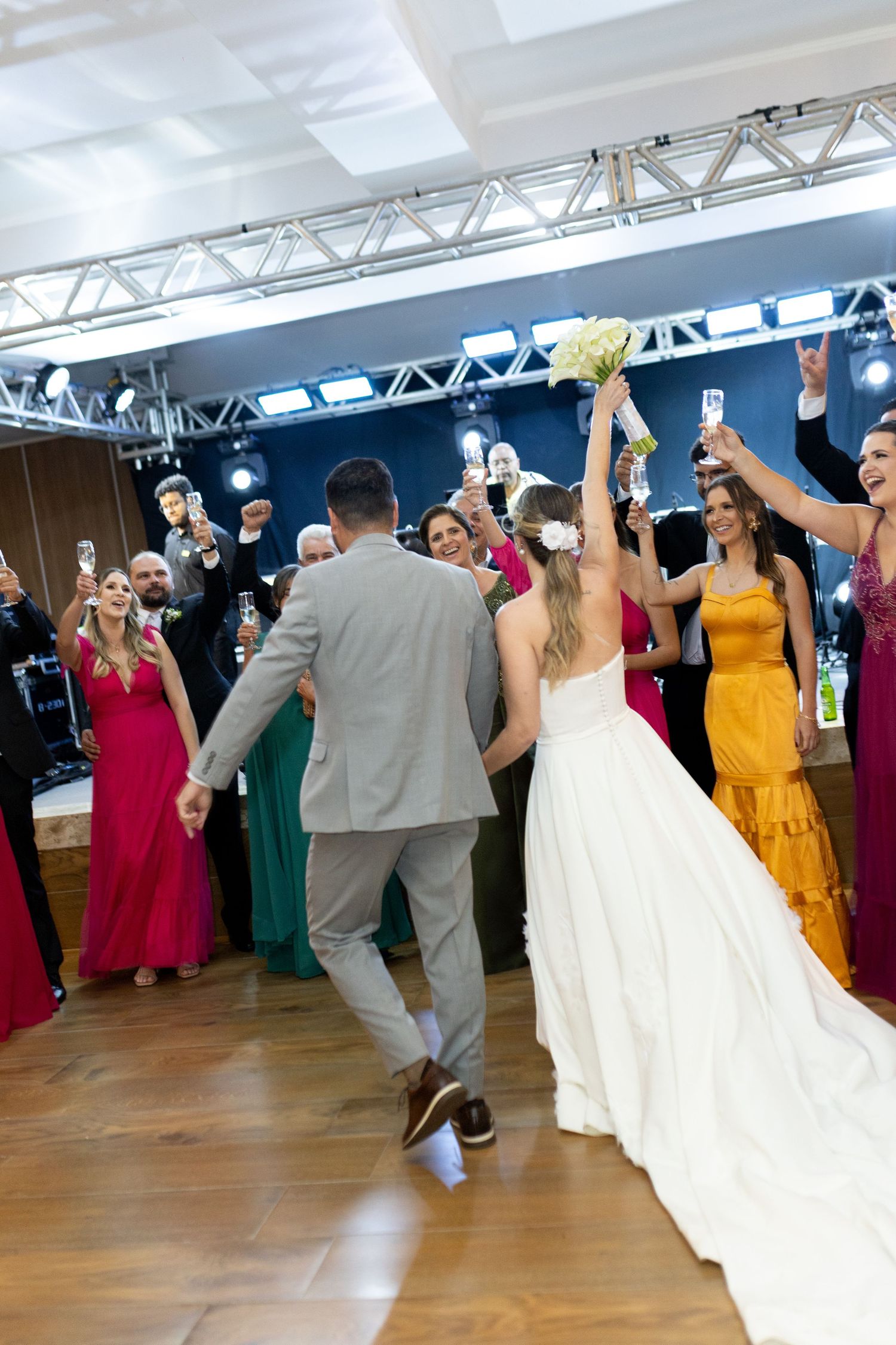 Bride and groom dancing joyfully at wedding reception surrounded by guests in colorful formal attire on wooden dance floor.