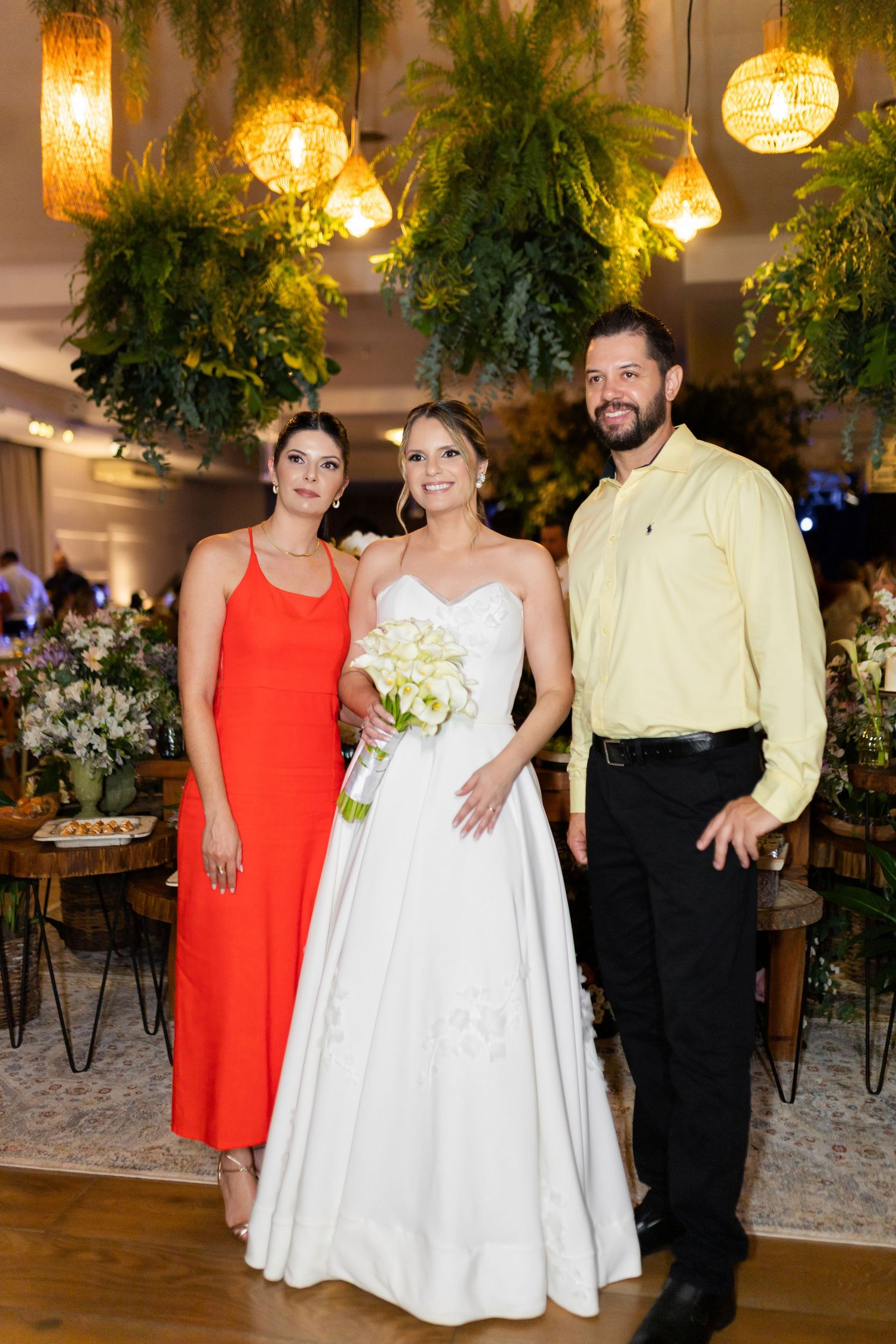 Bride in white gown holds bouquet flanked by woman in red dress and man in yellow shirt at elegant wedding reception.