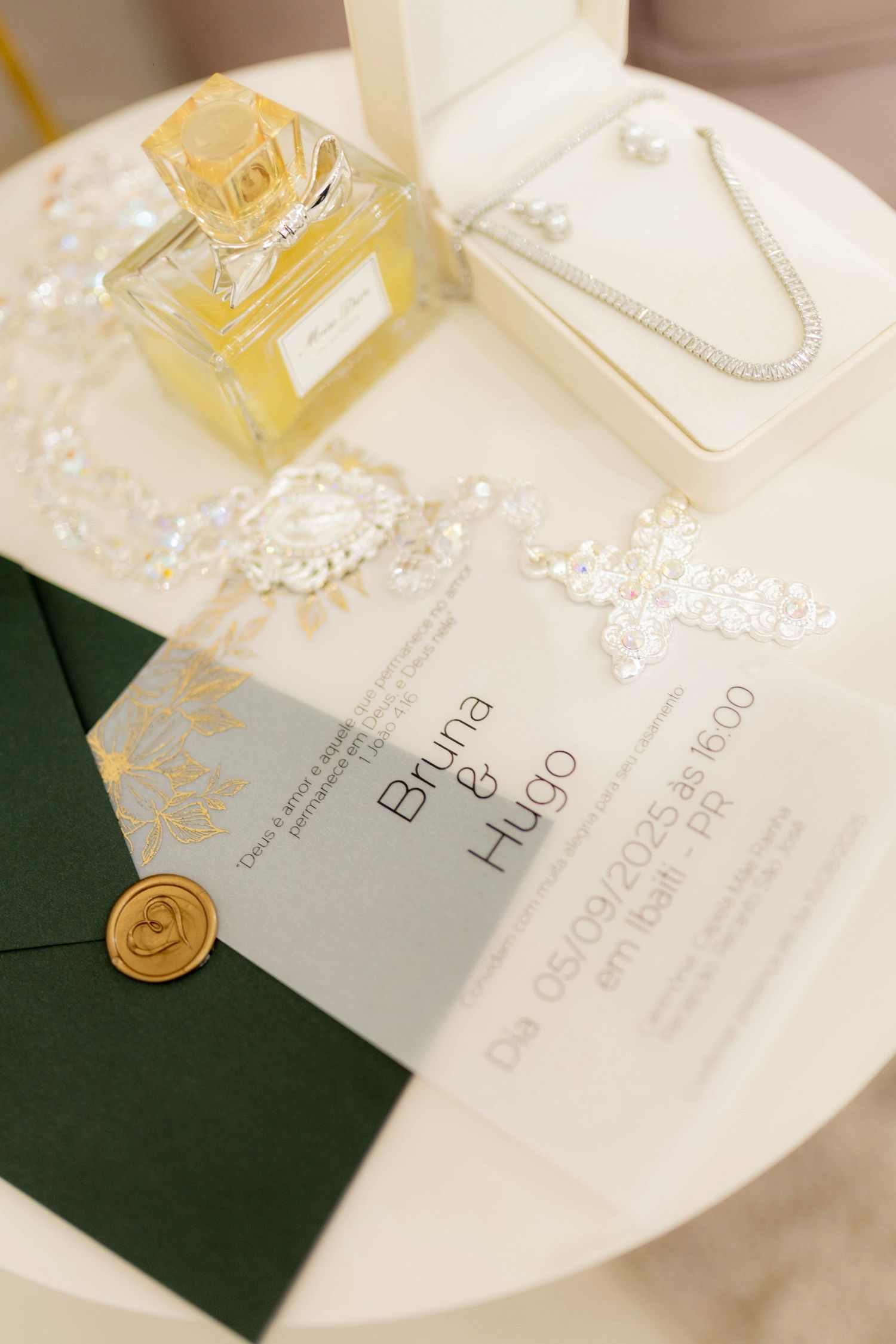Elegant wedding invitation for Bruna and Hugo with gold wax seal, green envelope, perfume bottle, and pearl necklace on white table.